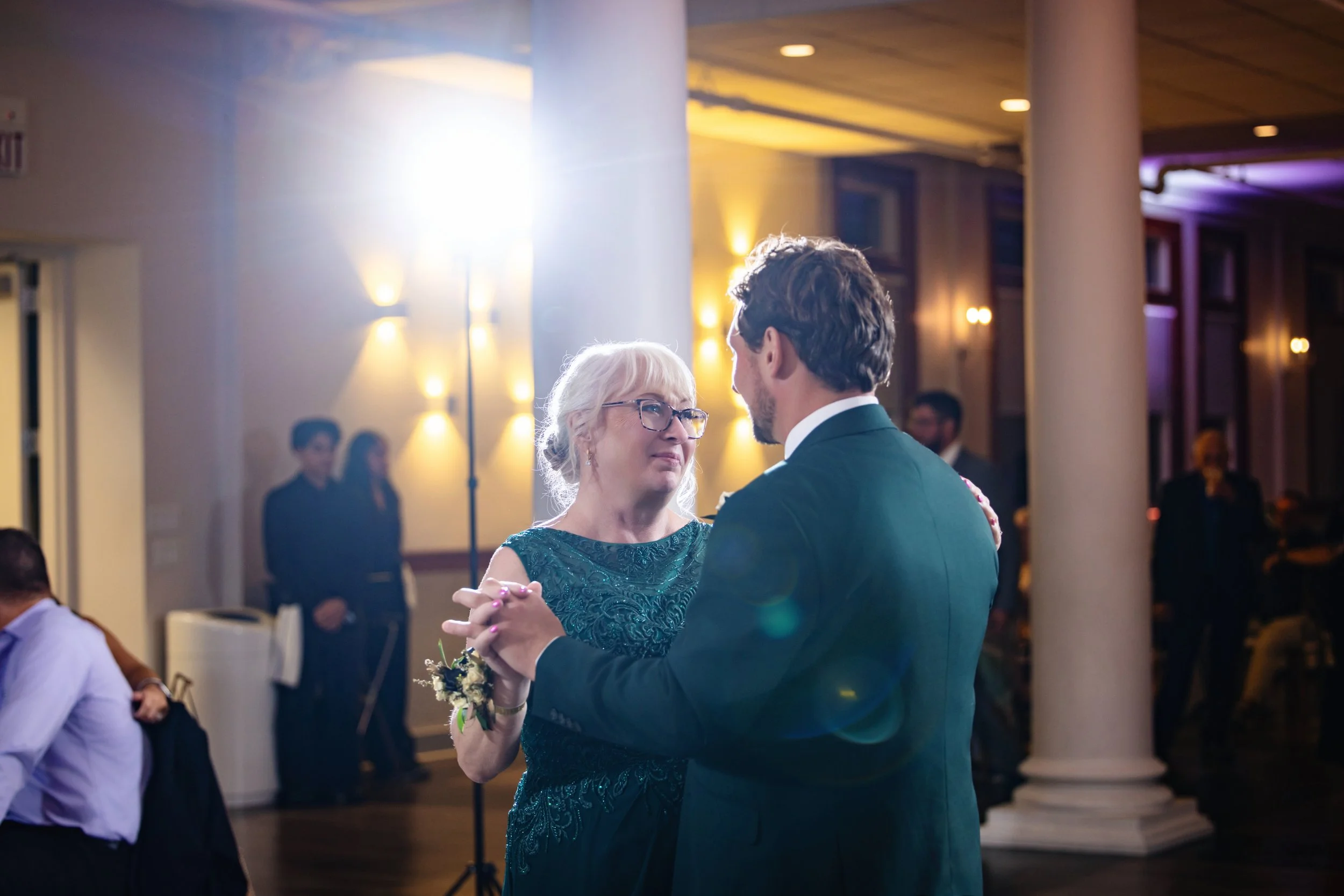 A woman and a man are dancing together at a formal event, with the woman wearing a green lace dress and the man in a dark suit. They are holding hands and looking at each other, surrounded by other guests, with warm lighting and elegant decor in the 