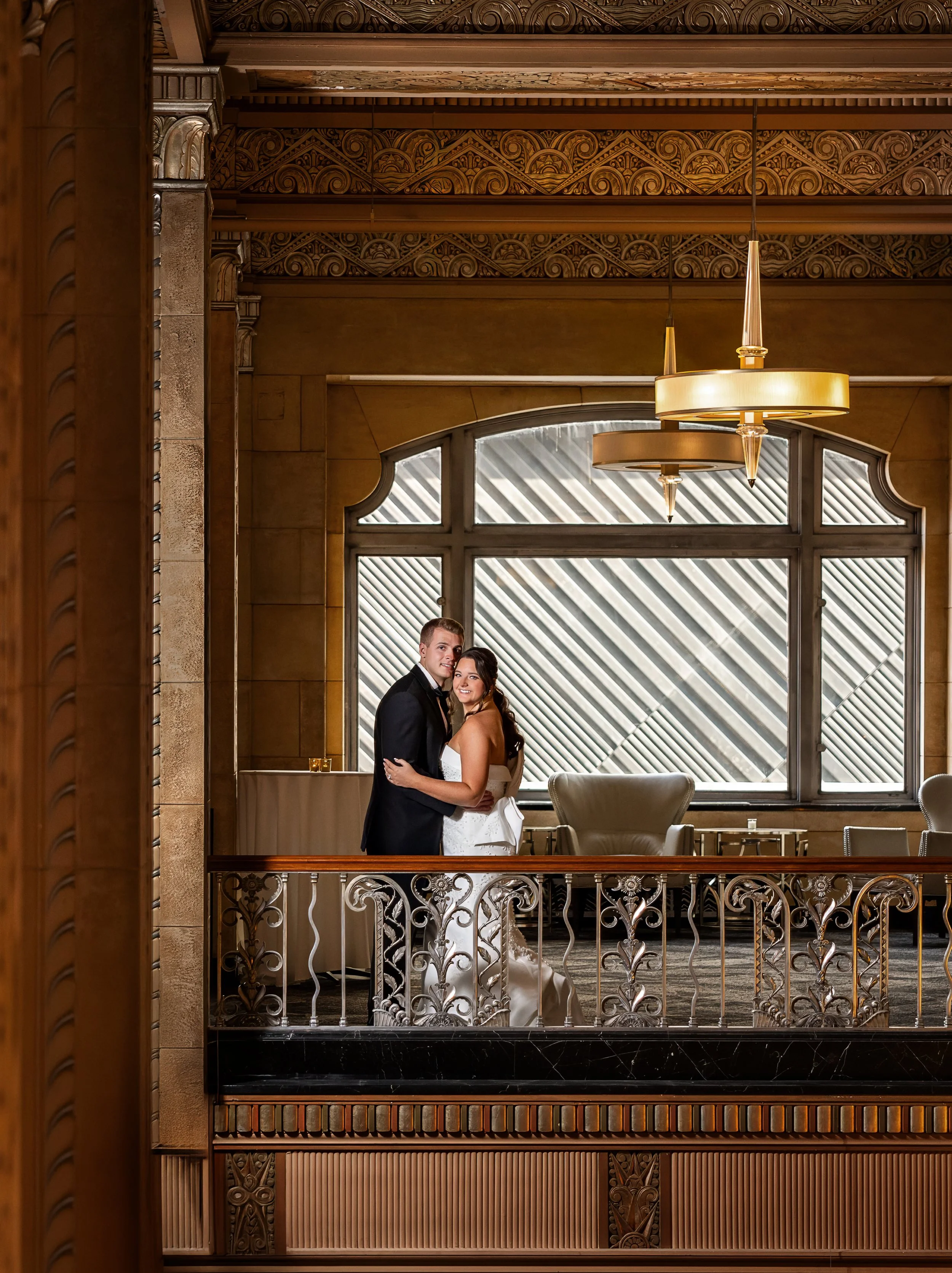 A bride and groom standing close together inside a luxurious building with ornate woodwork and large windows.