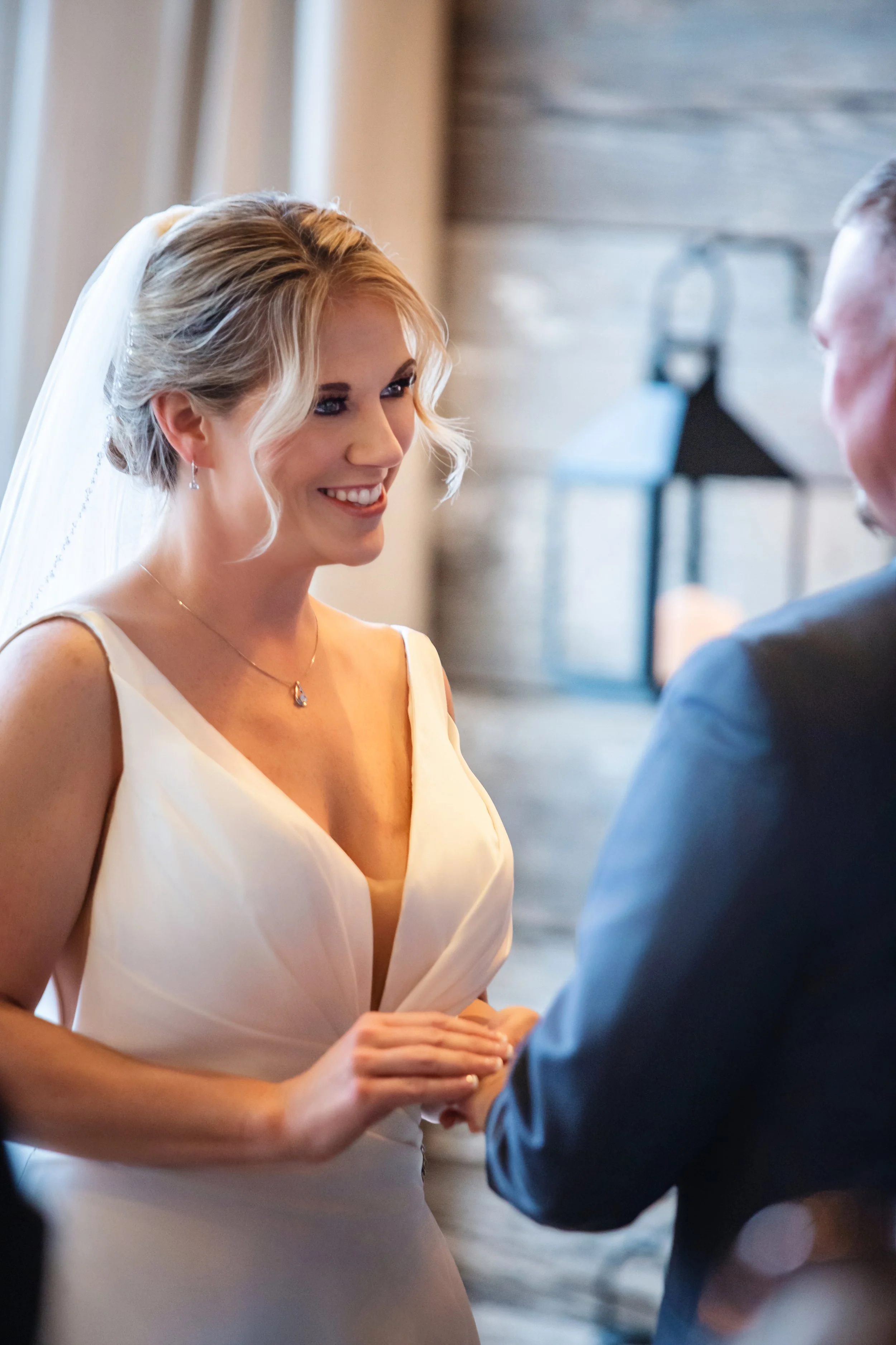 A bride with blonde hair styled in loose waves, wearing a white wedding dress and a veil, smiling while holding hands with a groom dressed in a dark suit.
