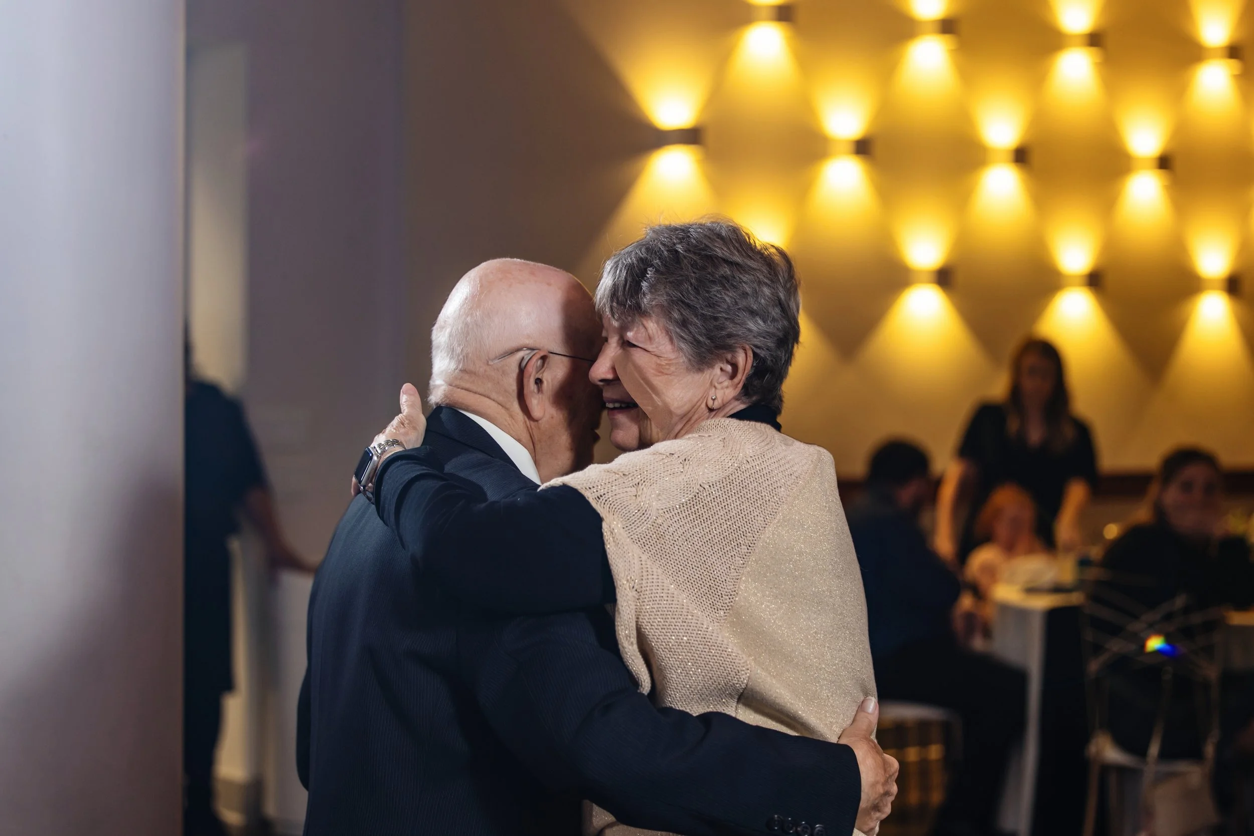An elderly man and woman dancing closely together with smiles in a warmly lit room, with blurred people sitting at tables in the background.