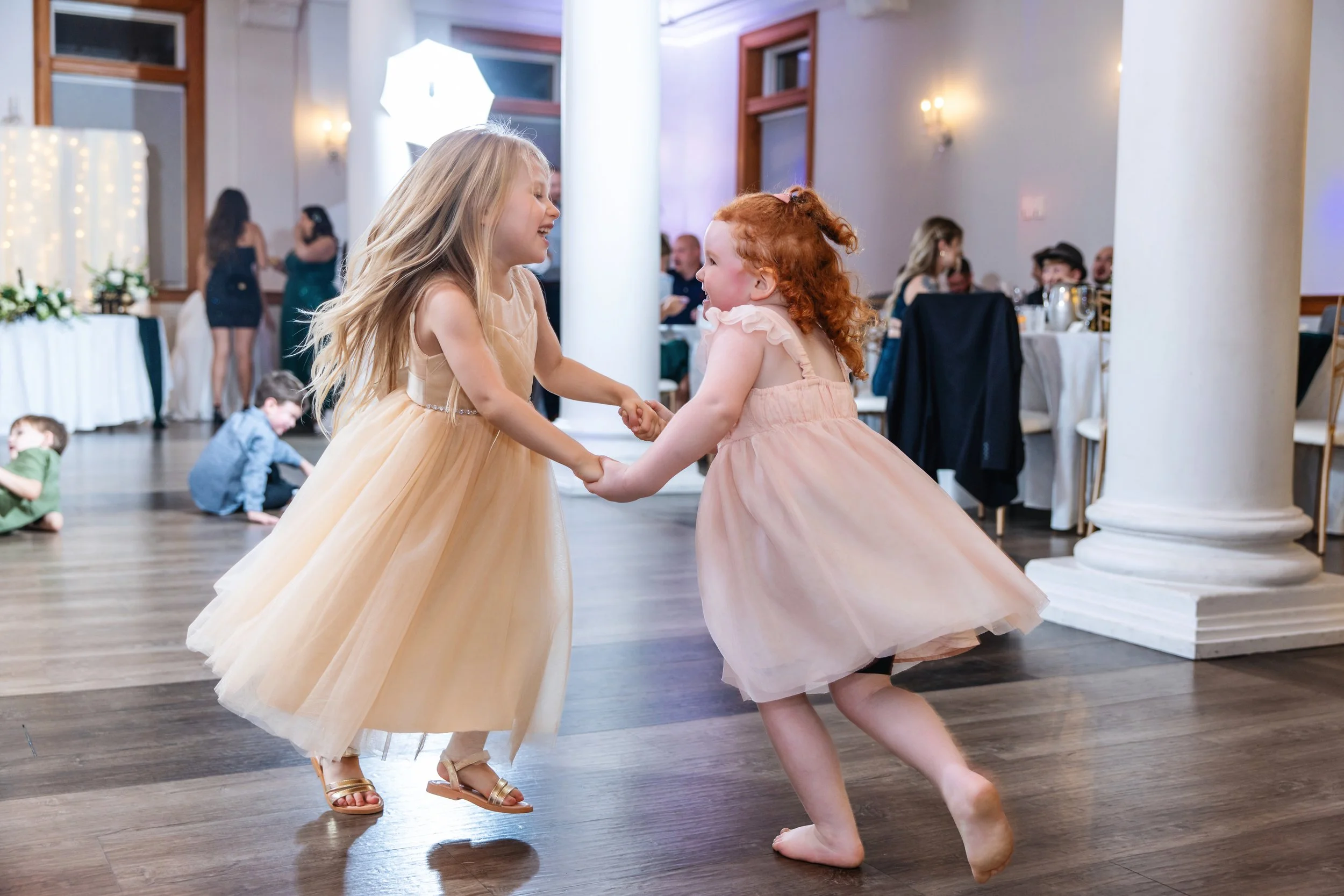 Two young girls dancing together at a party or celebration. One girl with long blonde hair in a light-colored dress and sandals, and the other girl with curly red hair in a pink dress, holding hands and spinning.