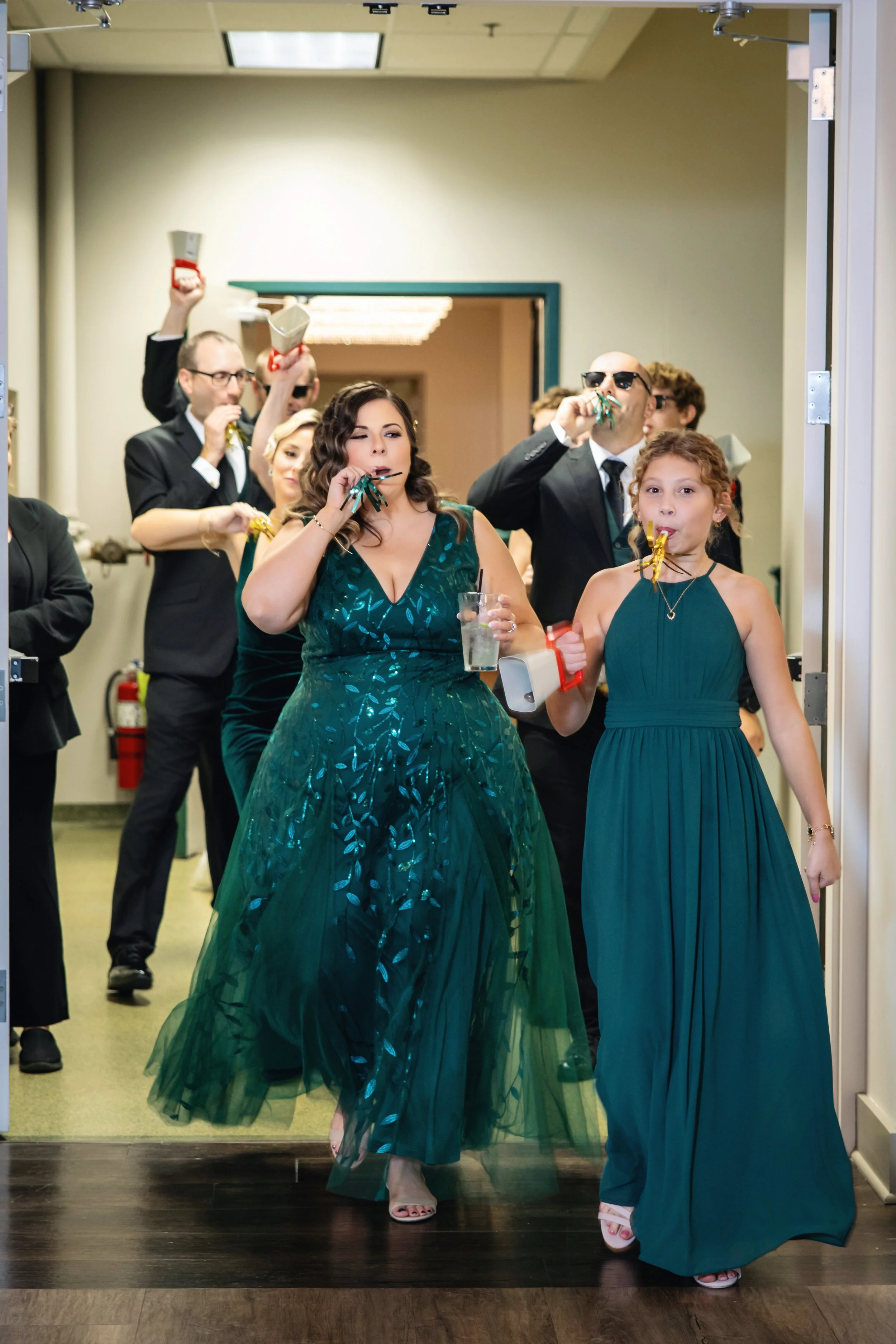 Group of people dressed in formal attire celebrating, some wearing sunglasses, blowing party horns, and holding noisemakers, as they walk through a doorway at an indoor event.