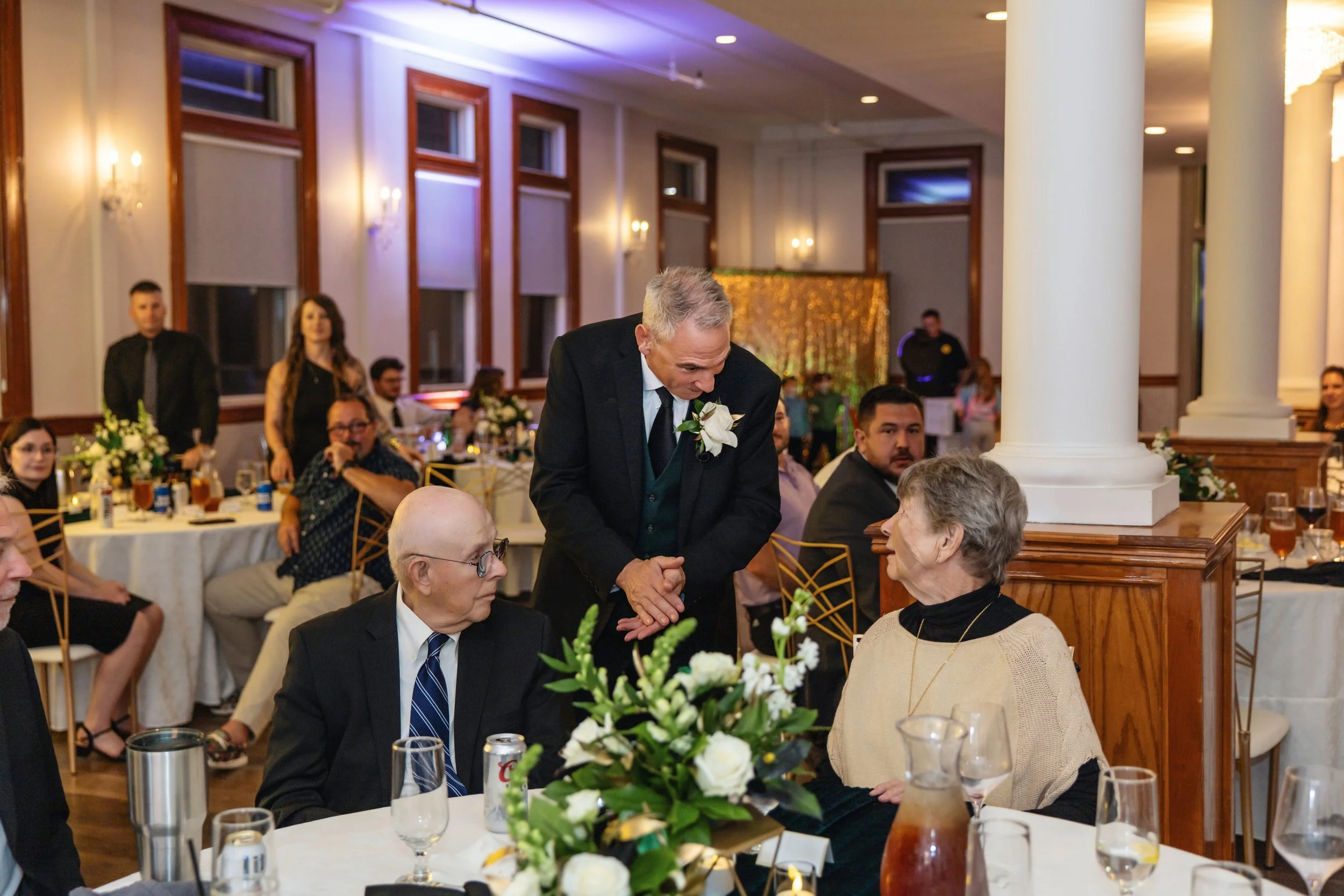 A wedding reception in a banquet hall with guests seated at tables decorated with white flowers. An older man with gray hair, wearing a dark suit and a white boutonniere, is shaking hands with an elderly woman. Other guests are visible in the backgro