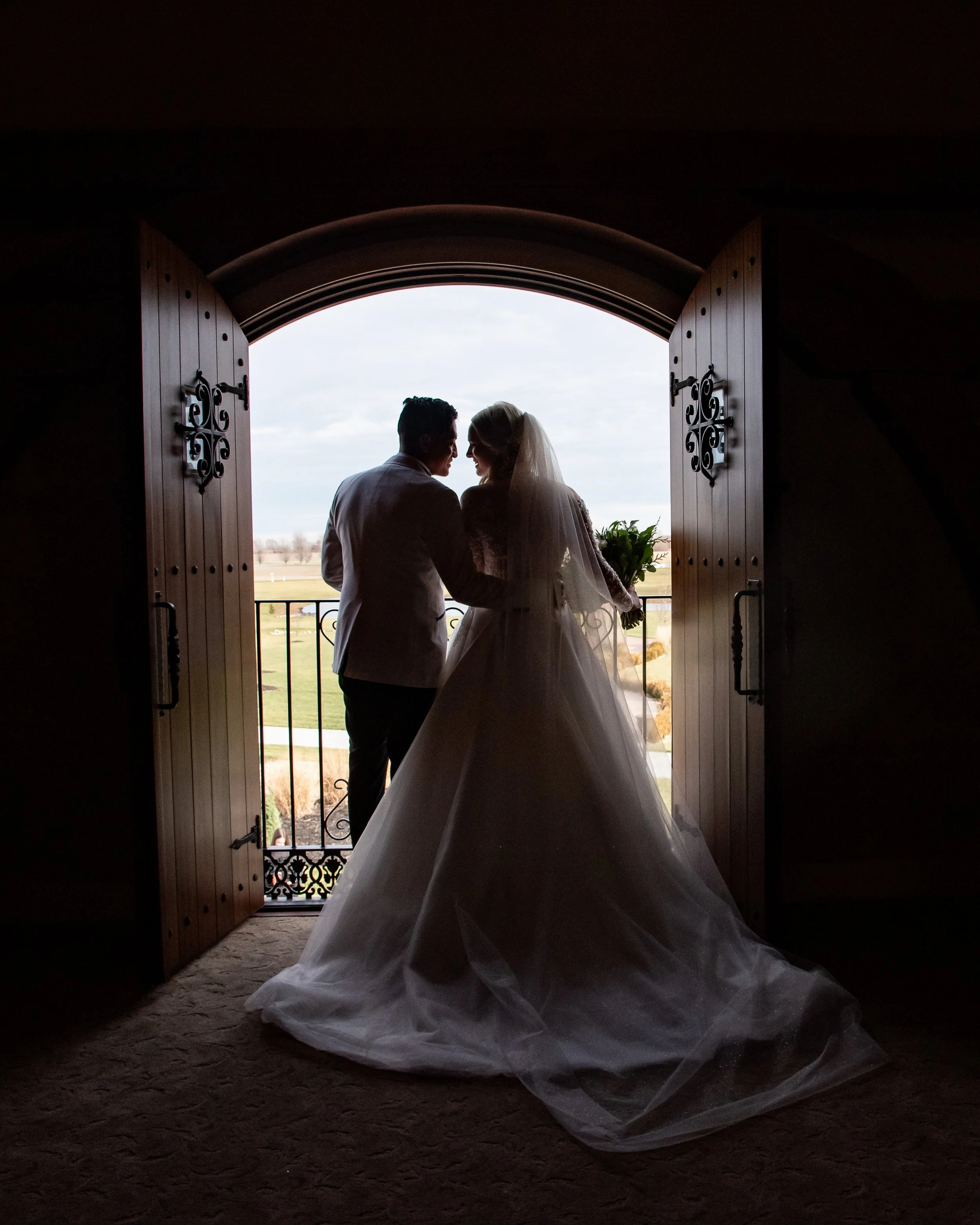 A bride and groom standing closely together under a large open doorway leading outside, with the bride holding a bouquet and wearing a veil.