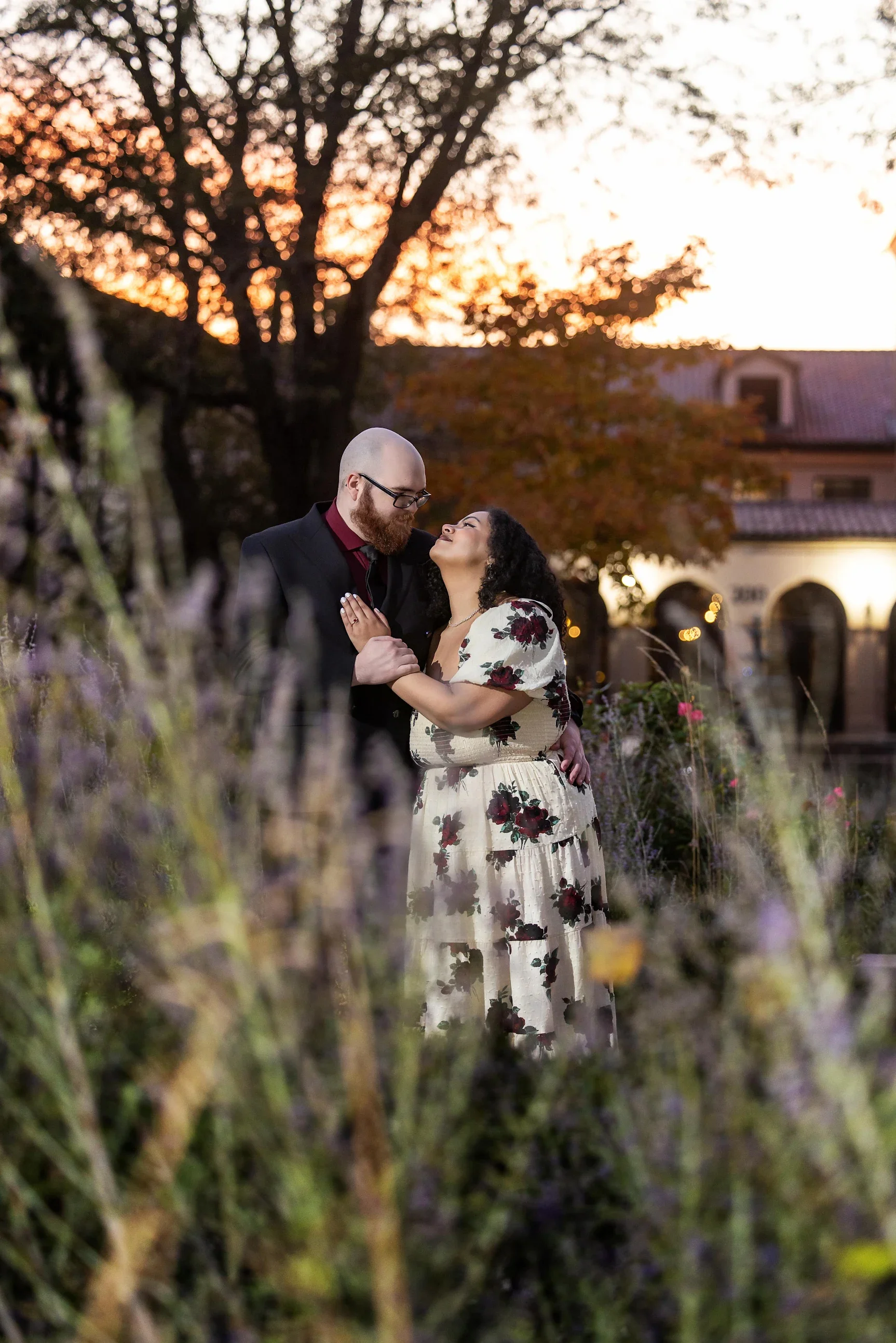 A couple embracing outdoors during sunset with a tree and buildings in the background.