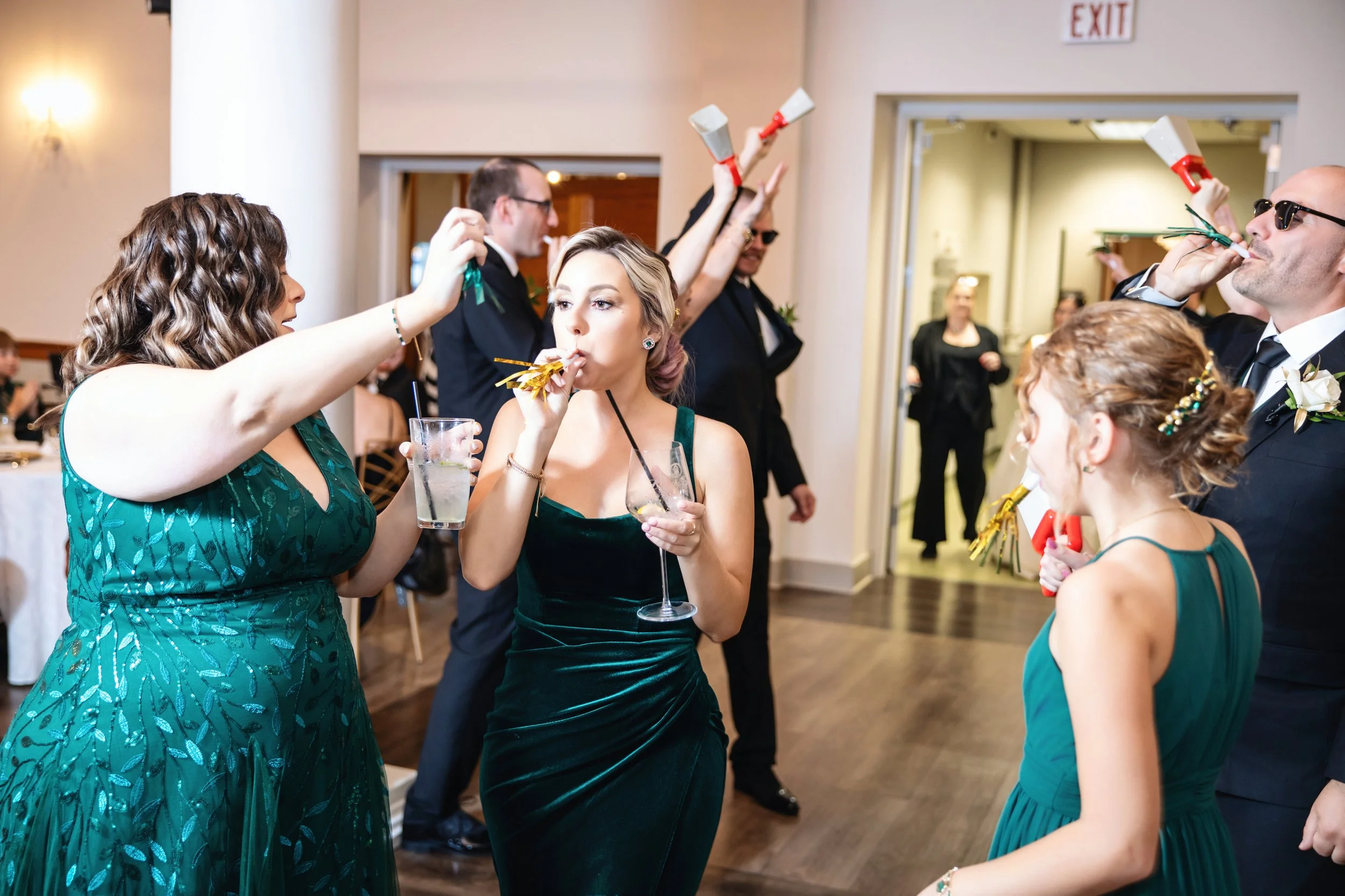 People dressed in formal attire at a celebration, with some blowing party noisemakers and holding drinks, in a decorated indoor venue.