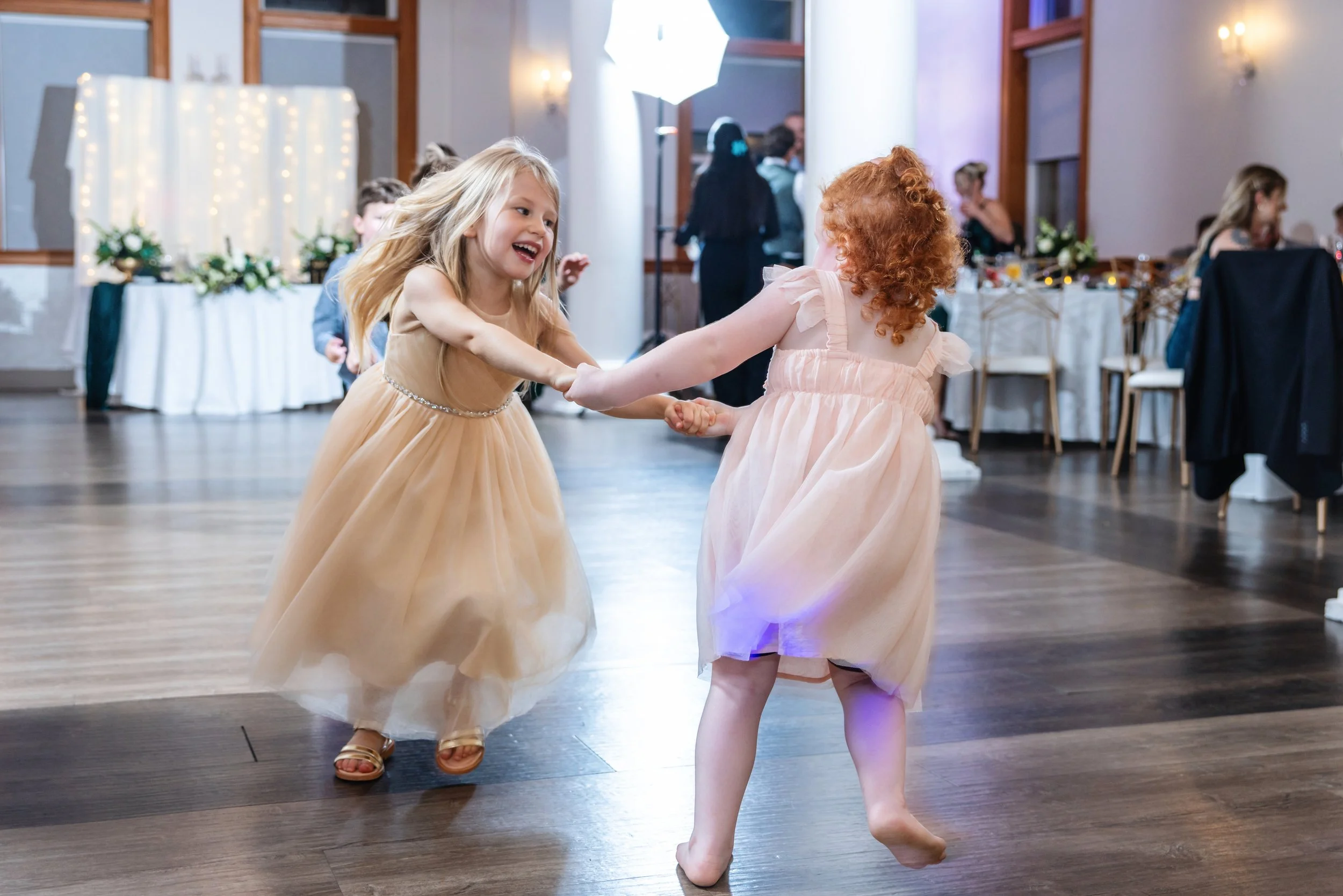 Two young girls dancing and holding hands at a party or celebration, wearing long, light-colored dresses, in a decorated banquet hall.