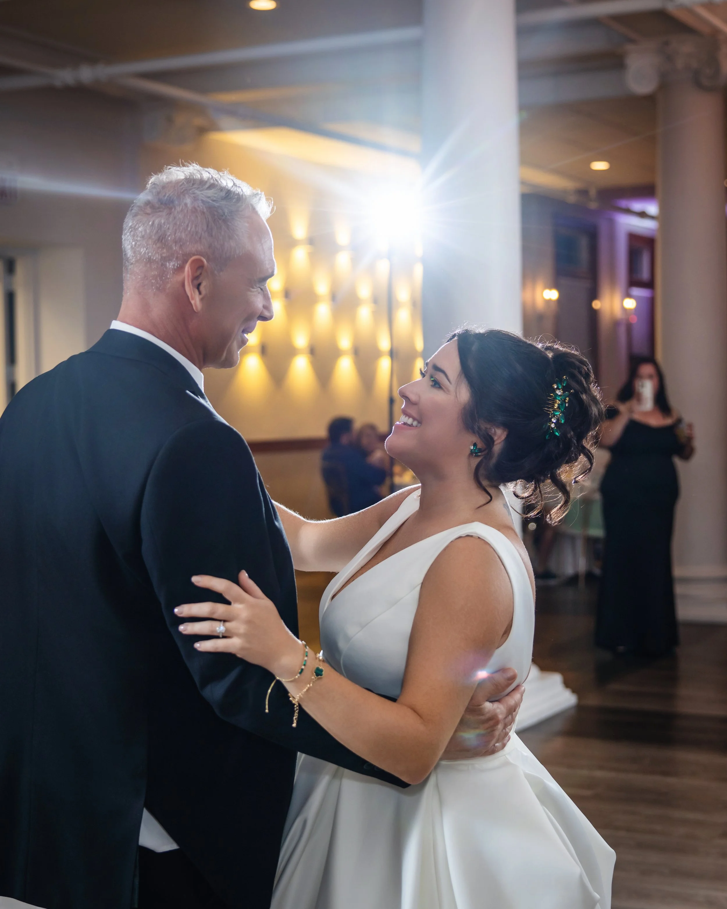 A couple dances closely together at a wedding reception, with the man wearing a dark suit and the woman in a white gown. The woman looks up at the man with a smile, and the background features warm lighting and other guests.