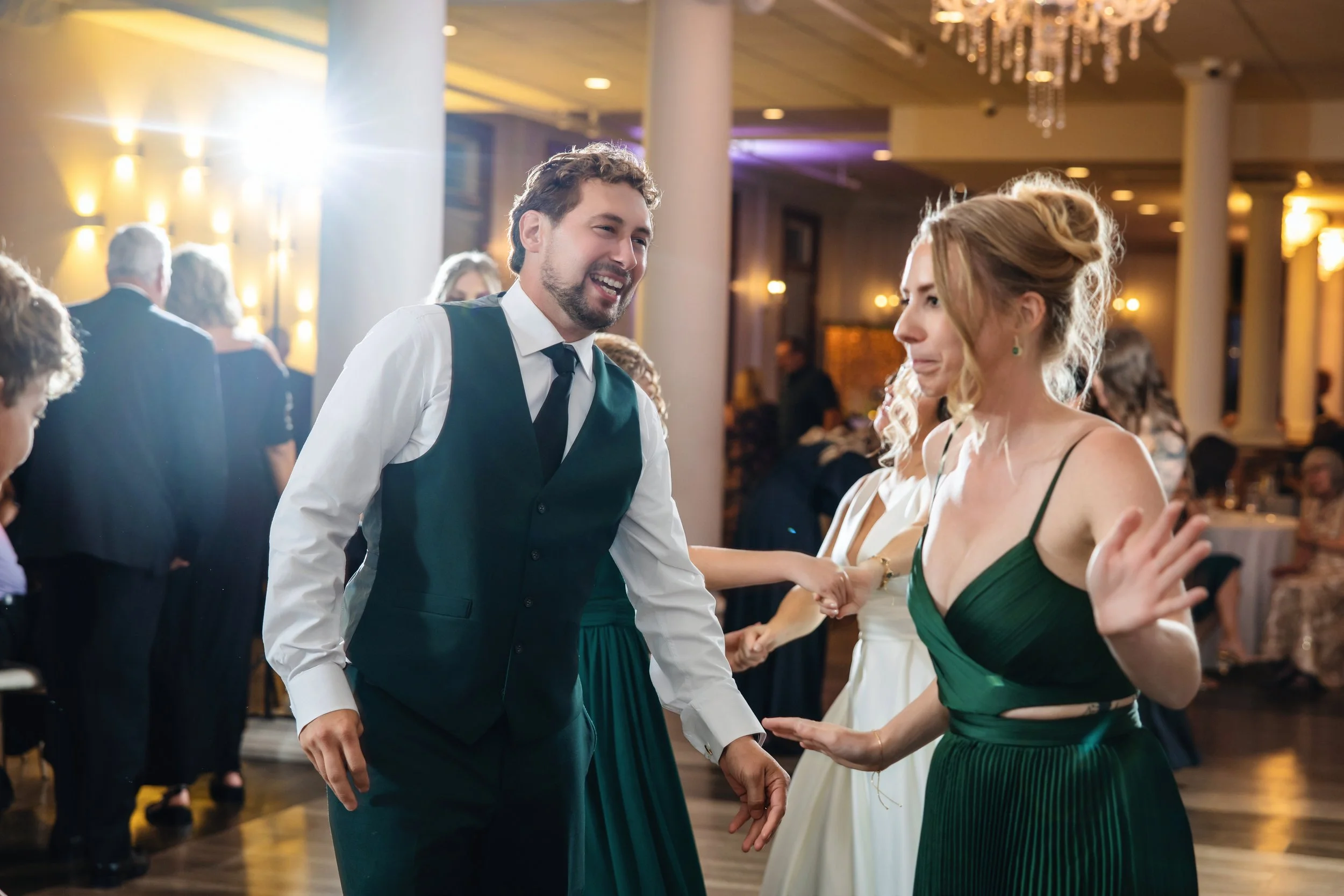Couple holding hands and dancing at a formal event or wedding reception, with guests and a chandelier in the background.