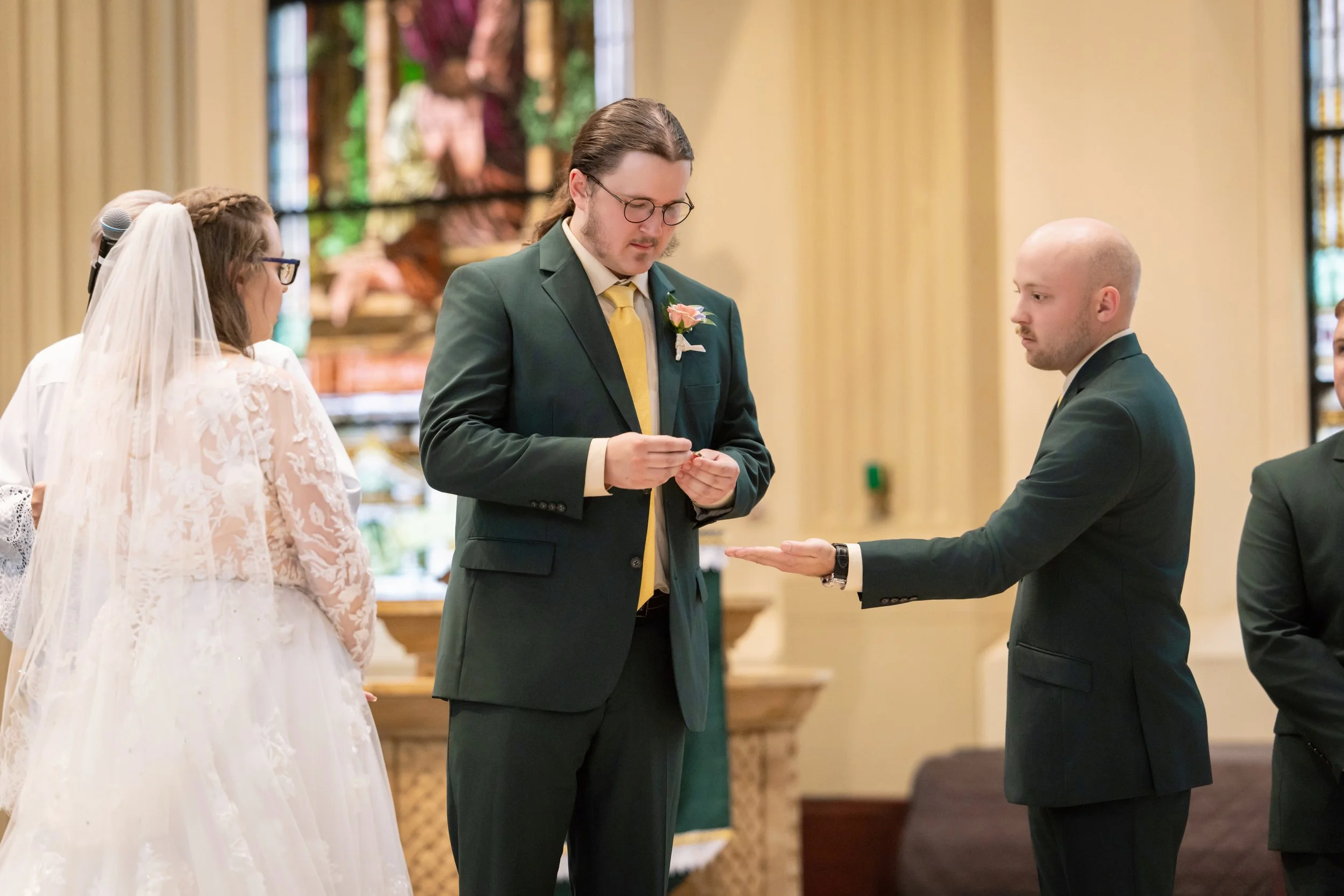 A wedding ceremony with a bride, groom, and two witnesses. The bride is wearing a white lace wedding dress and veil, the groom is dressed in a dark suit, and one witness is a man with long hair and glasses wearing a green suit with a yellow tie, hold