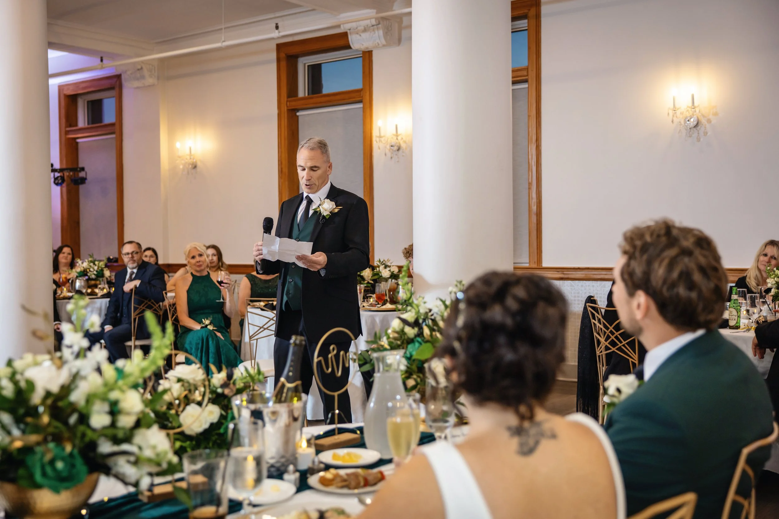 A man in a tuxedo giving a speech at a wedding reception, surrounded by seated guests at decorated tables with flowers and drinks.