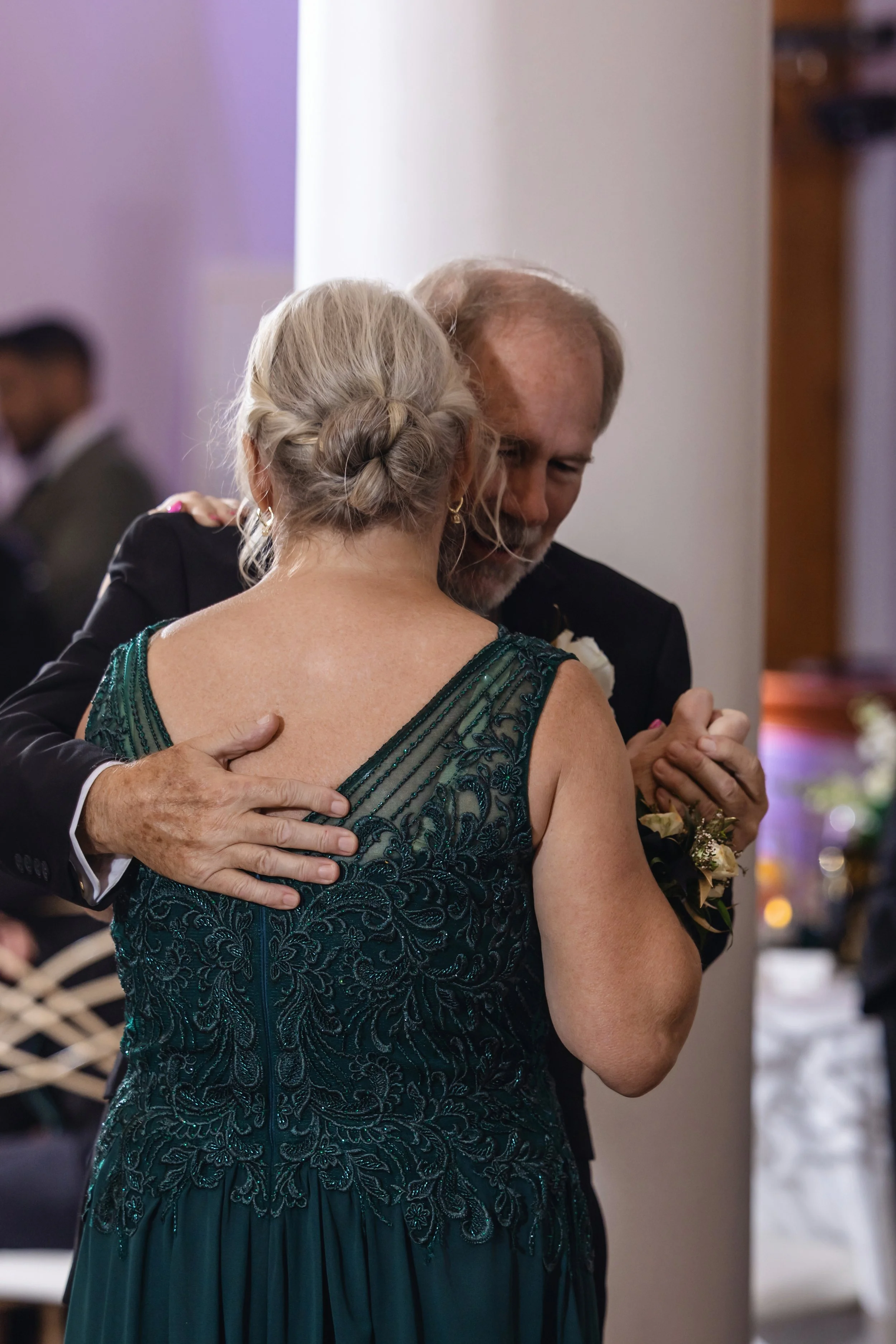 An elderly woman with gray hair in an updo and a man are dancing closely at a formal event, with other guests in the background.