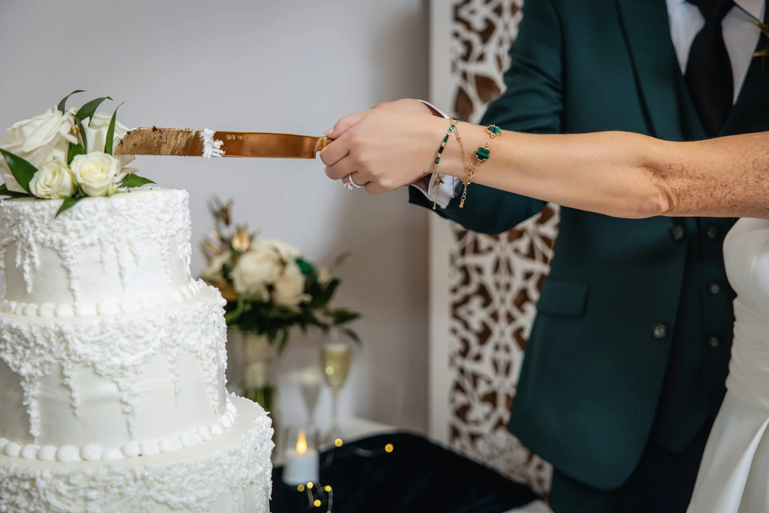 A person cutting into a white wedding cake decorated with white roses and greenery. The person is wearing jewelry on her wrist and a dark formal attire. The background has an ornate pattern and blurred floral arrangements.