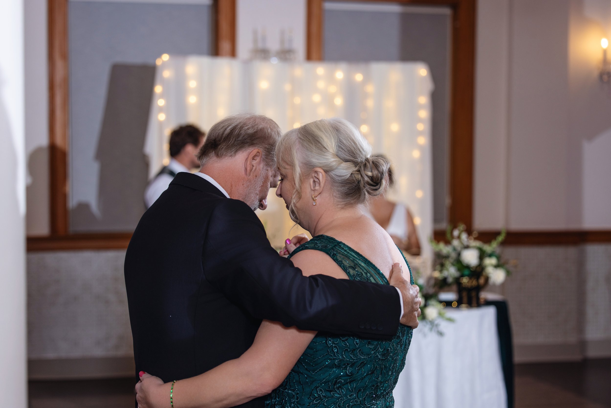 An elderly couple dancing closely at a wedding reception, with string lights and a wedding cake in the background.