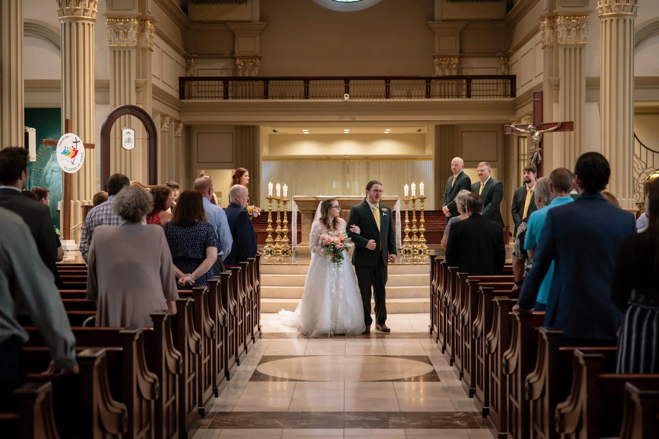 A bride and groom standing at the altar during a wedding ceremony in a church, with wedding guests seated on either side of the aisle, an ornate altar with lit candles, cross, and religious statues in the background.