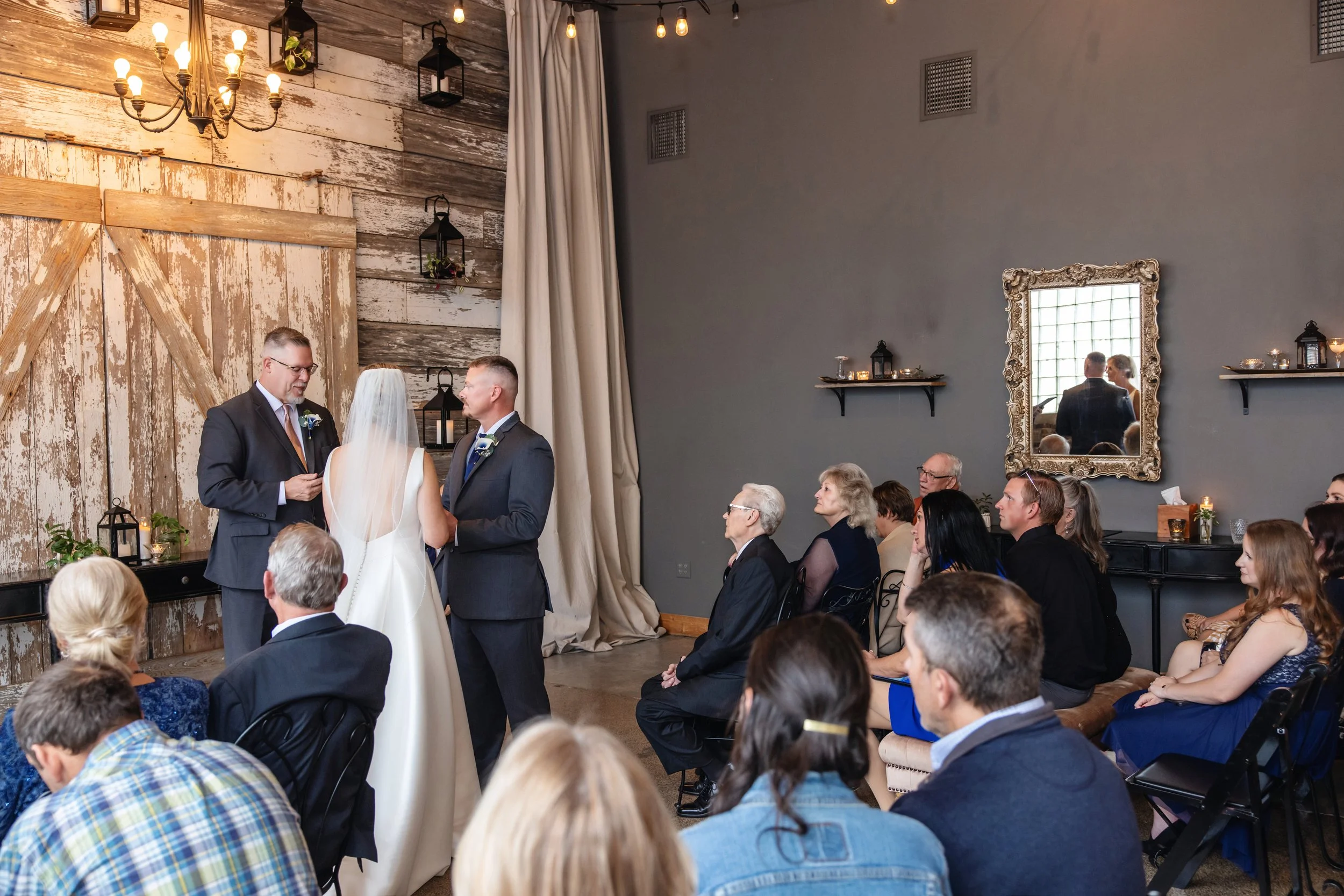 A wedding ceremony with the officiant, bride, and groom standing in front of guests seated indoors. The room has rustic decor with wooden panels and hanging lanterns. A large ornate mirror is mounted on the wall, and candles are placed on shelves.