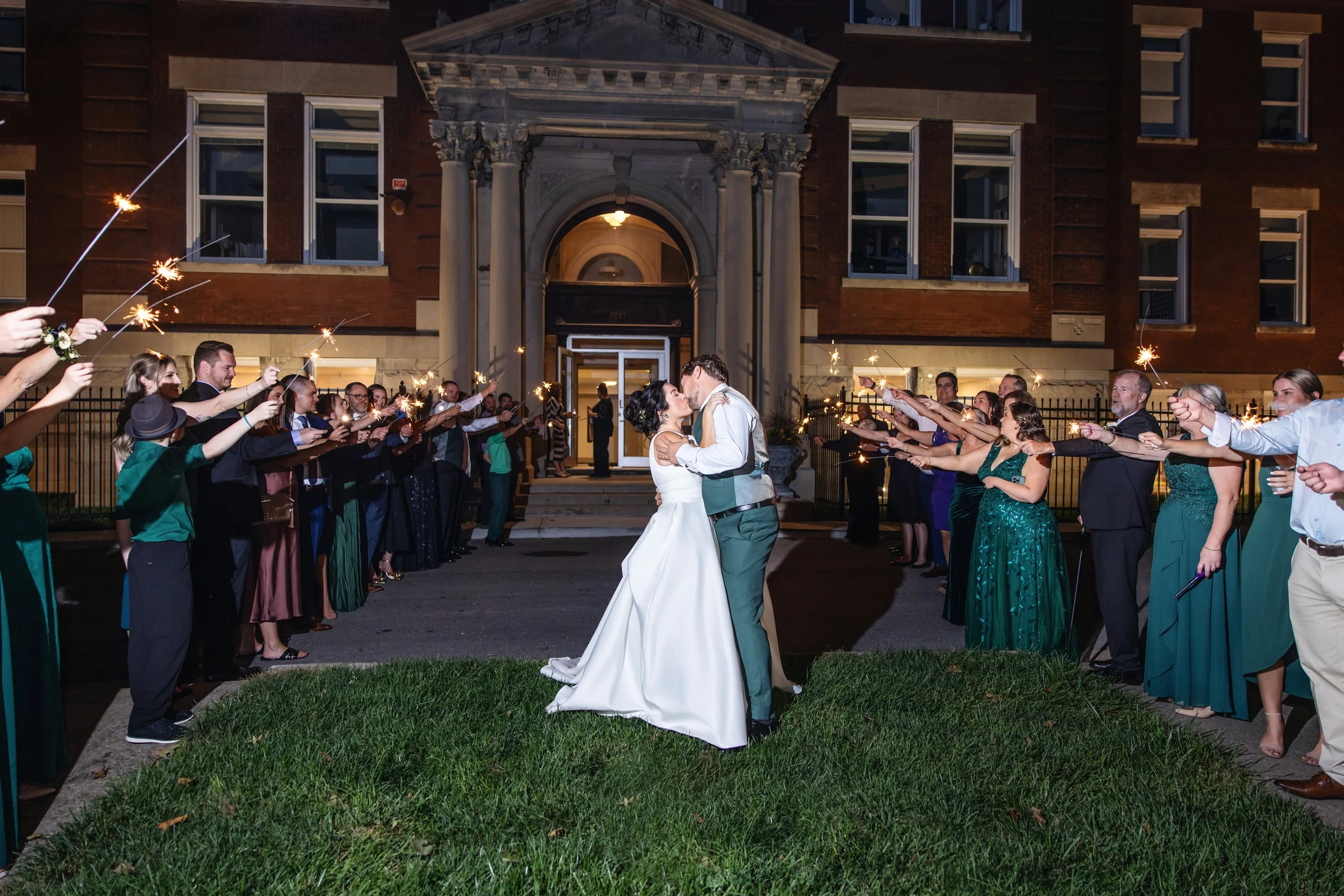 A bride and groom kissing in front of wedding guests holding sparklers outside a building at night.
