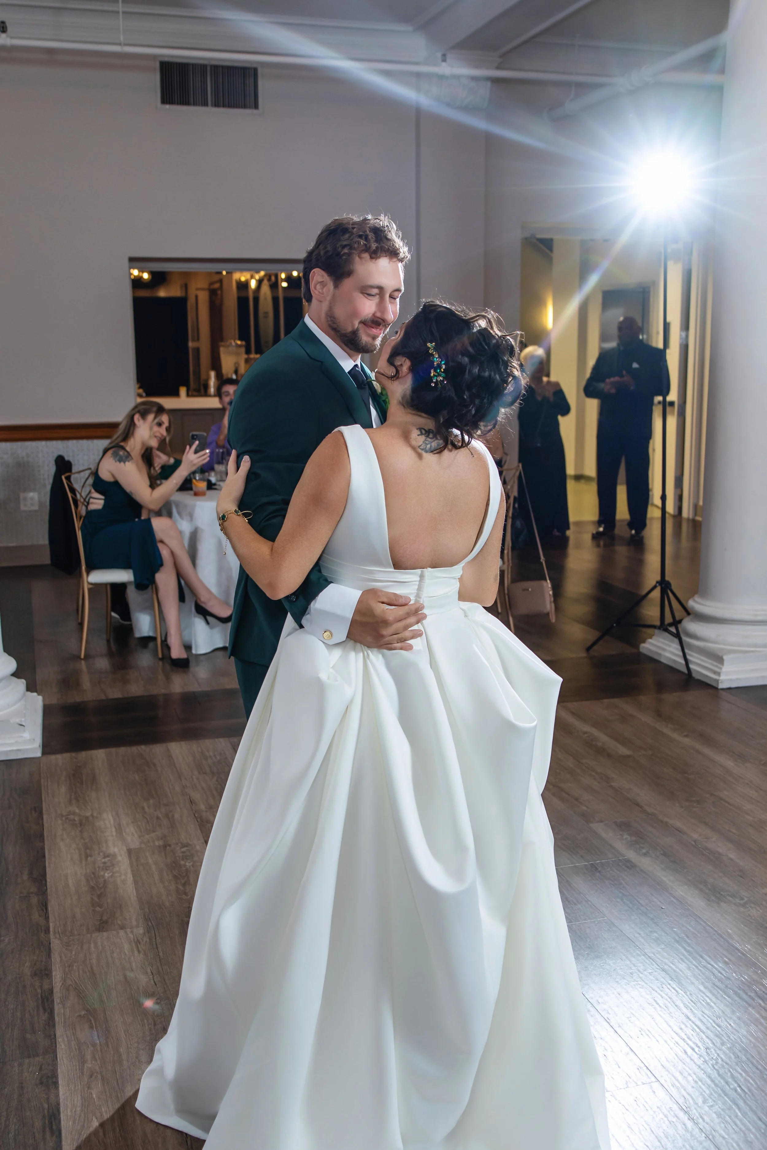 A bride and groom share a dance at their wedding reception. The bride is wearing a white gown with a full skirt, and the groom is wearing a dark suit. Guests are visible in the background, with some sitting at tables and others standing, watching the