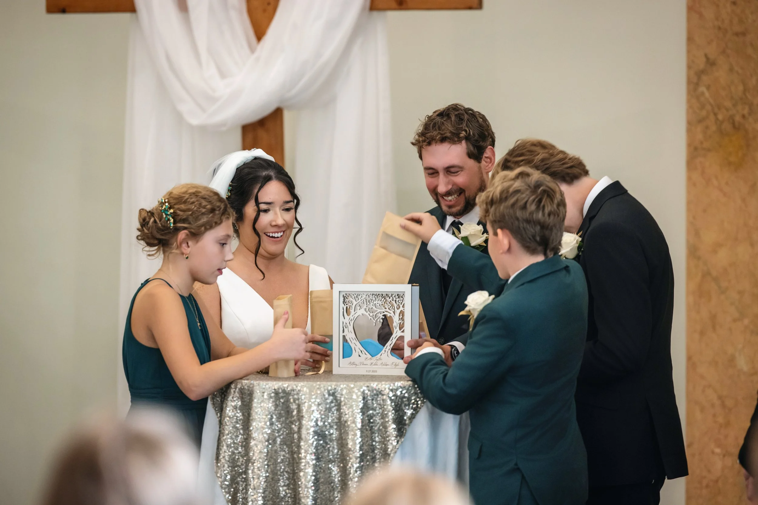A wedding scene with a bride in a white dress and a groom in a suit, surrounded by children, as they exchange gifts at a decorated table inside a church or reception hall.