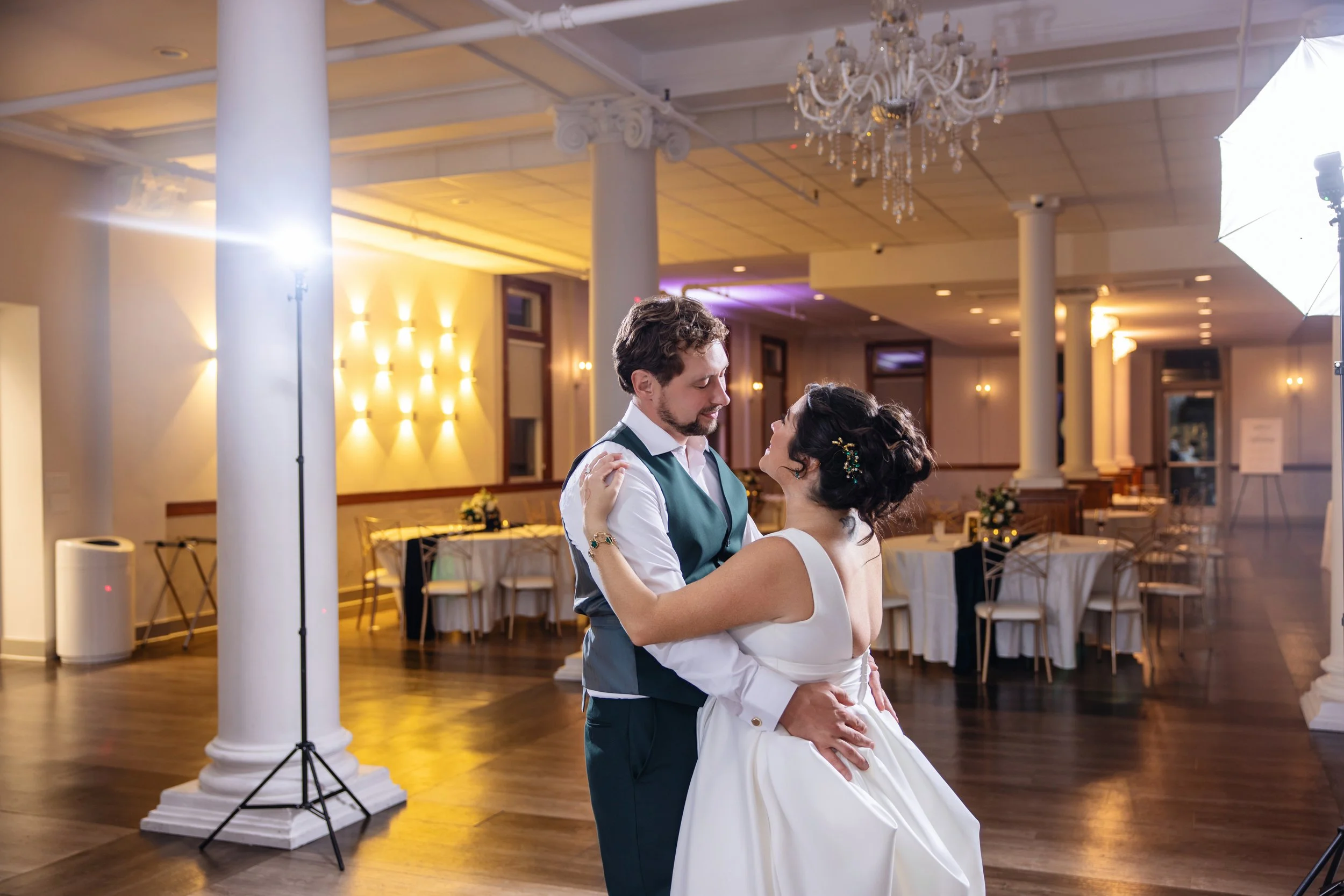 A couple shares a dance at their wedding reception in a decorated banquet hall, with elegant lighting and a chandelier overhead.