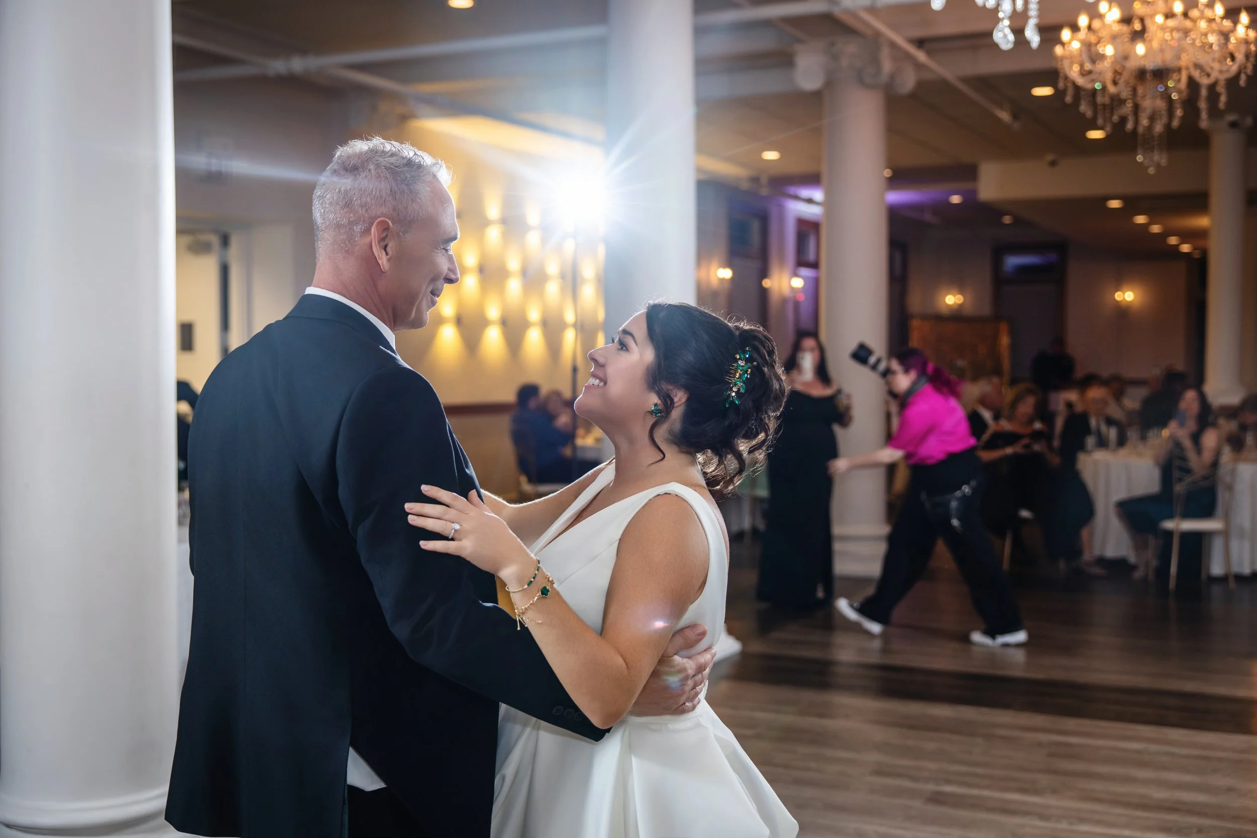 A bride and groom dancing at their wedding reception in a decorated banquet hall, with guests seated at tables in the background.