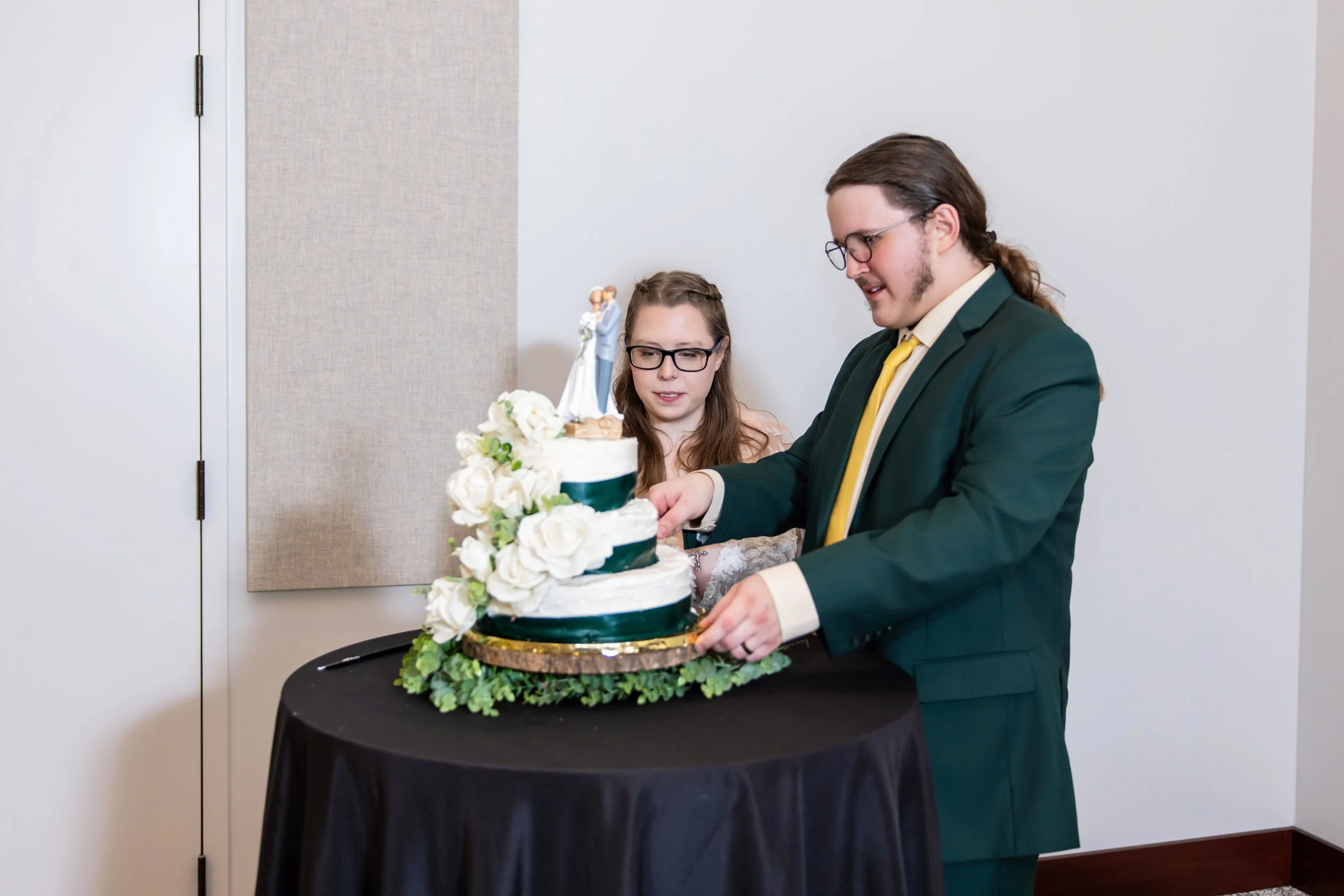 A man and woman, dressed there wedding attire, cutting a wedding cake decorated with white flowers and a bride and groom topper. They are standing at a black table with a black tablecloth, indoors with plain white and beige walls.