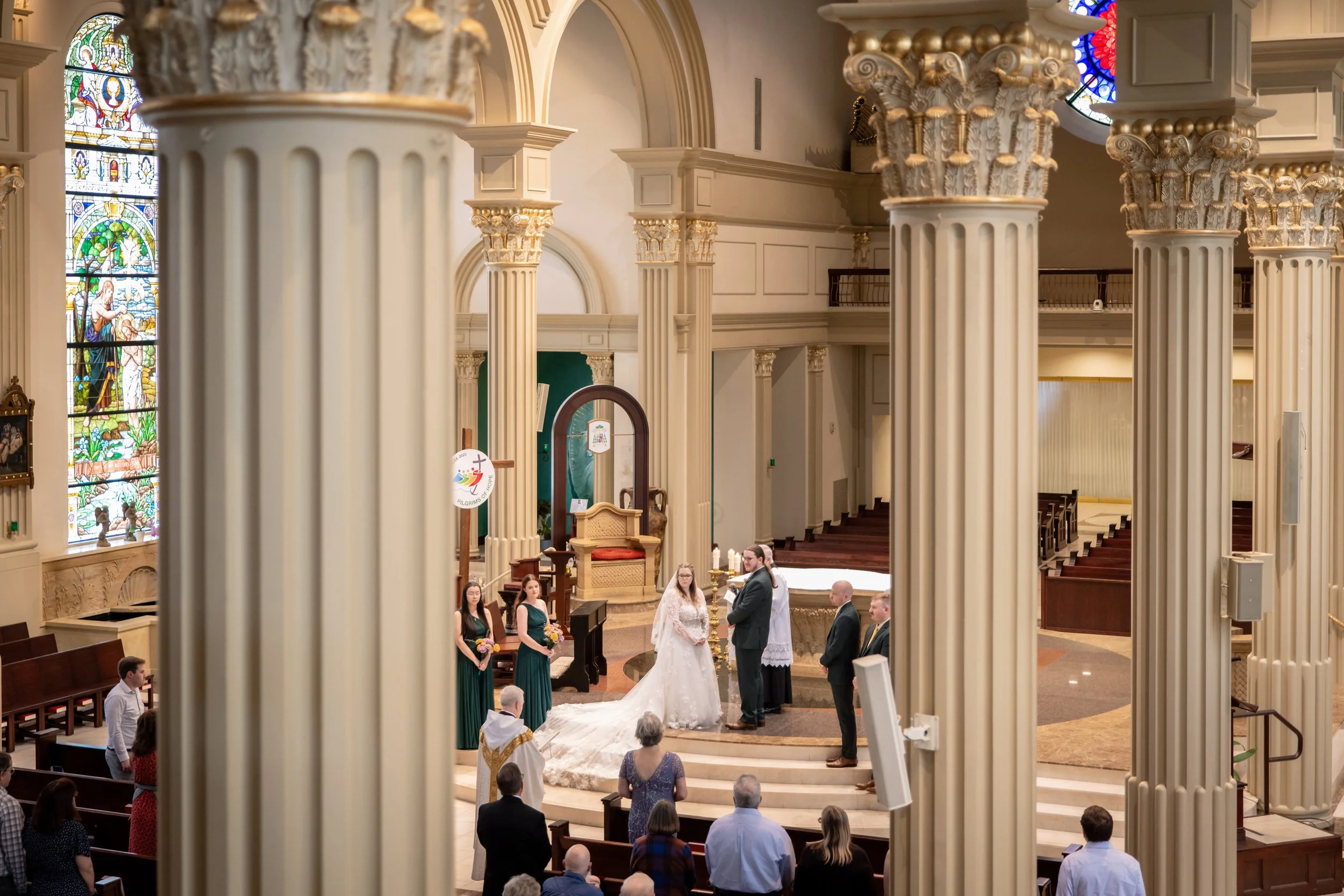 The image shows the interior of a church with wedding ceremony in progress. Columns with ornate capitals, stained glass windows, and pews are visible. A bride and groom are at the altar, surrounded by bridesmaids and officiants.