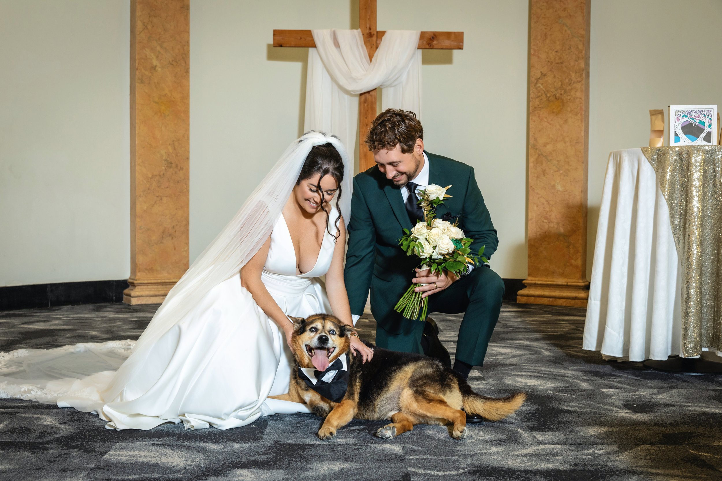 A bride and groom kneeling together at their wedding ceremony, smiling and petting a large, happy dog wearing a bow tie, in front of a cross draped with white fabric.