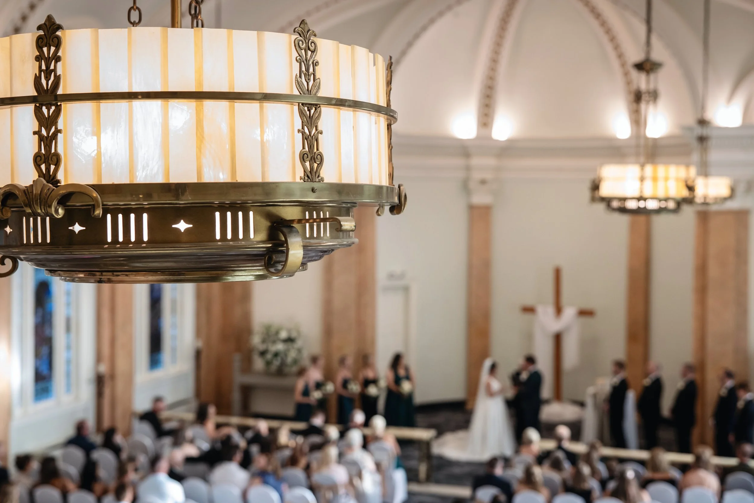 Decorative chandelier hanging in a church as people gather for a service, with a cross at the altar in the background.