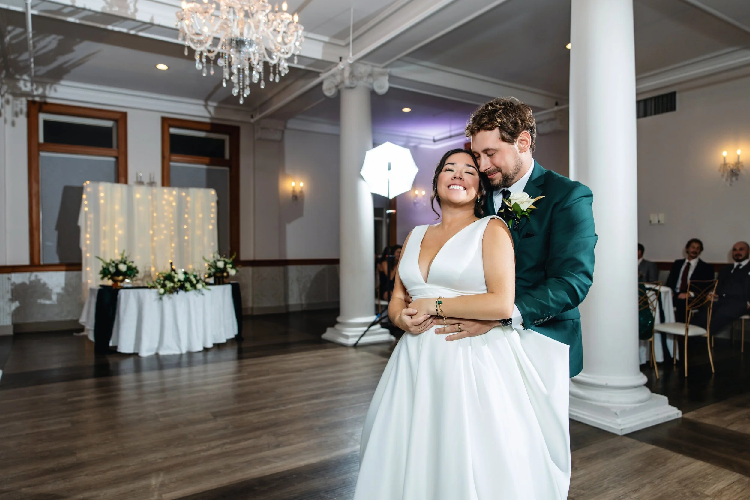A bride and groom share a moment during their wedding reception in an elegant, well-lit banquet hall decorated with chandeliers and floral arrangements.