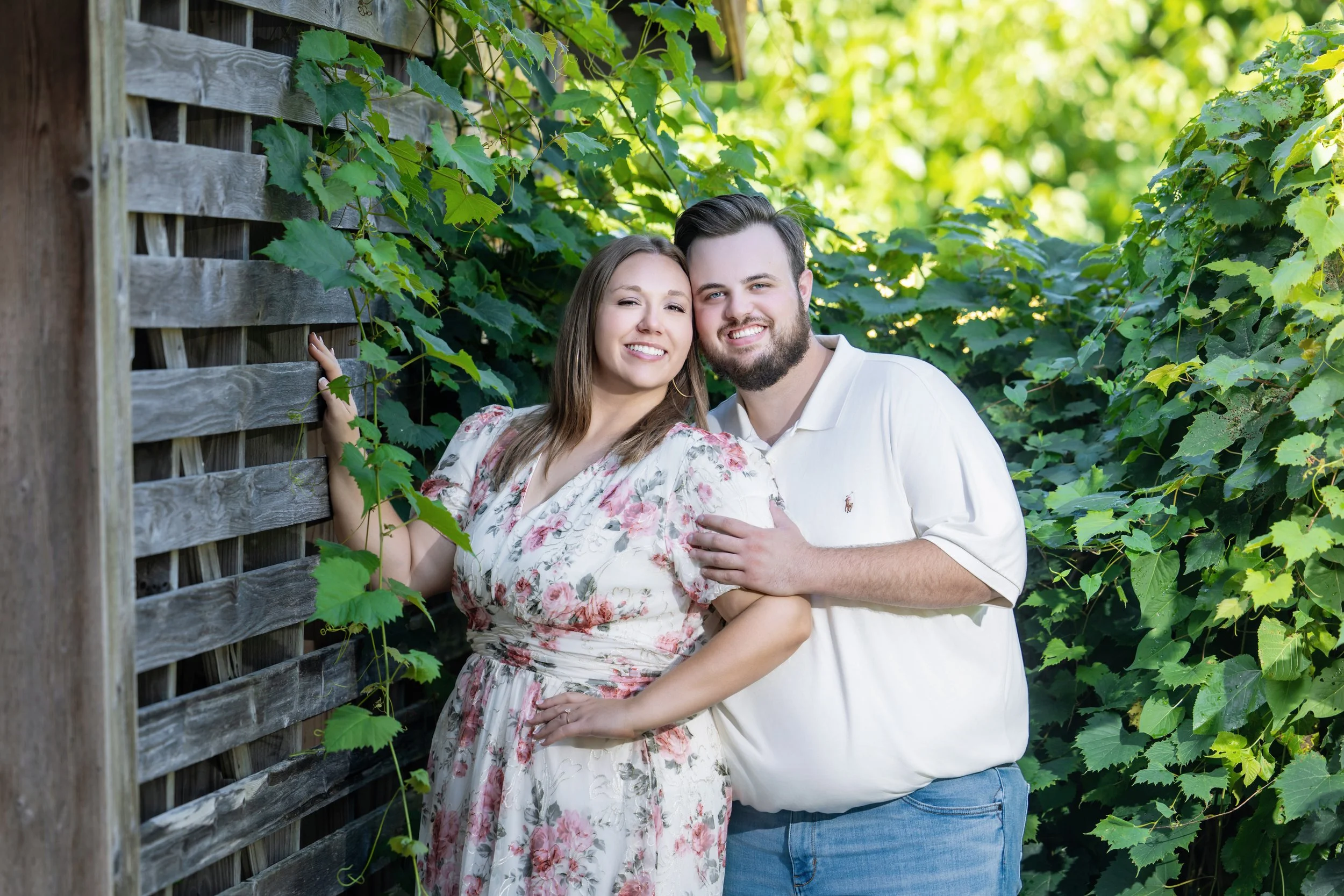 A smiling couple with the woman winking, standing beside a wooden fence overgrown with green vines and leaves in a lush garden.