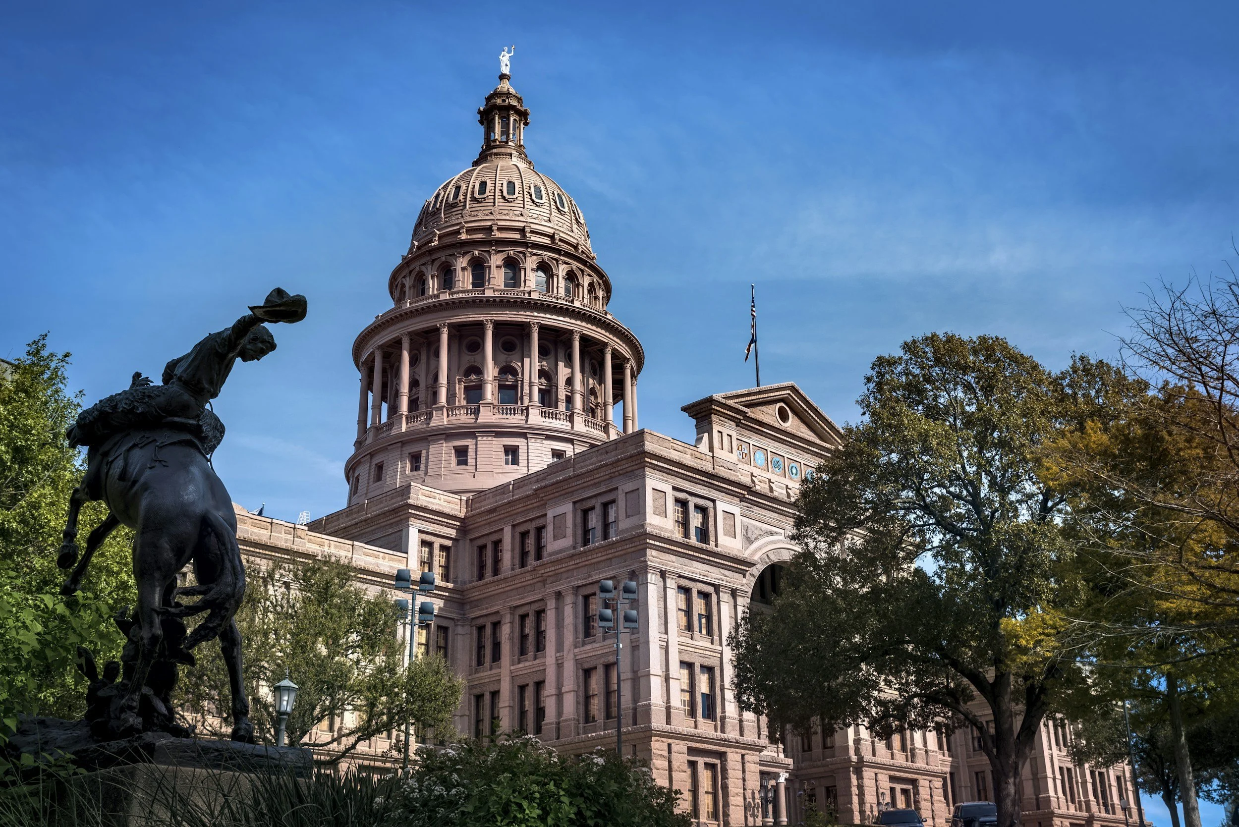 The Texas State Capitol building in Austin, Texas, under a clear blue sky, with trees partially obscuring the view and a statue of a horse and rider in the foreground.