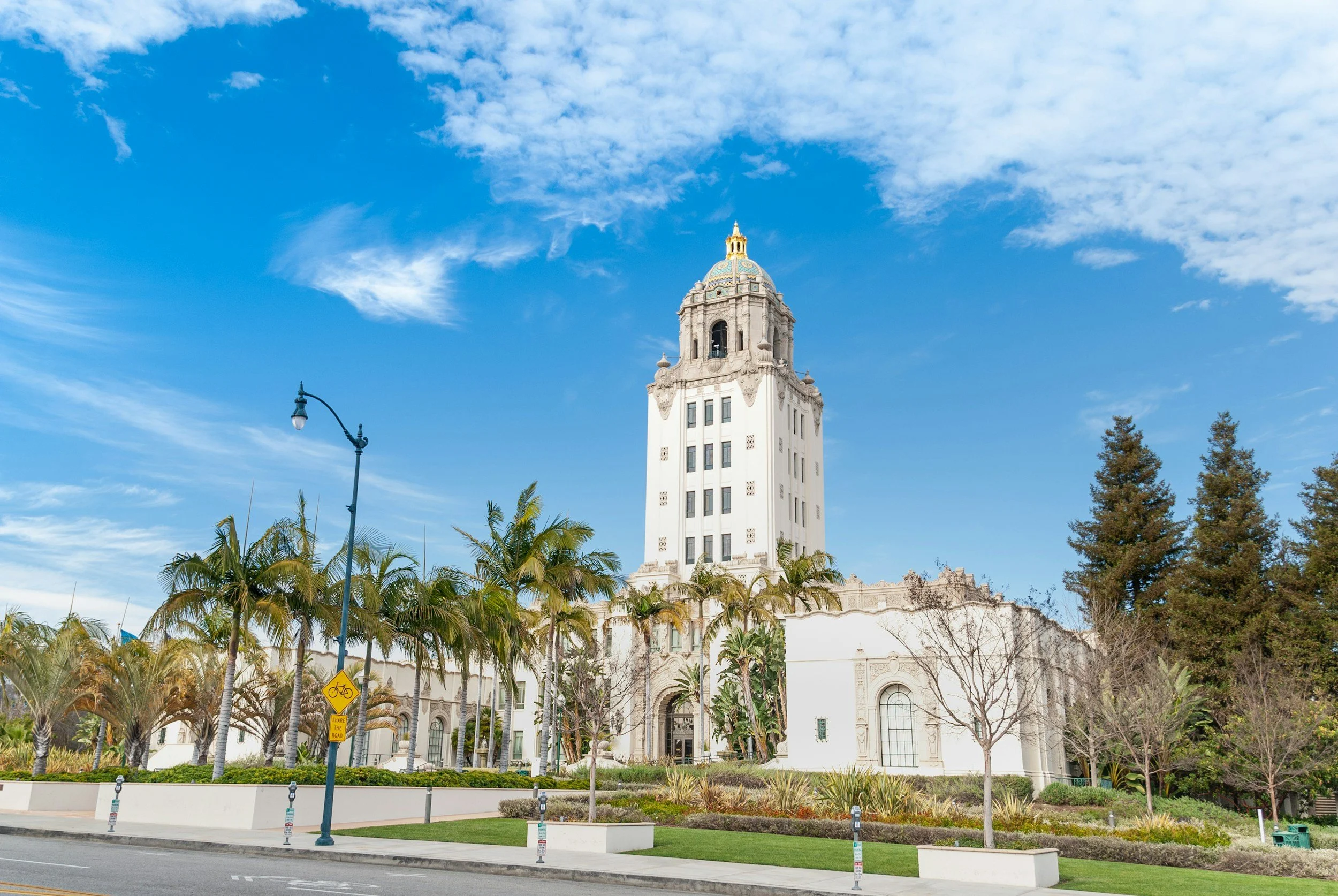 A historic white building with a tall clock tower topped with a decorative golden dome, set against a bright blue sky with scattered clouds. Palm trees and a street with a bicycle lane and street signs are in the foreground.