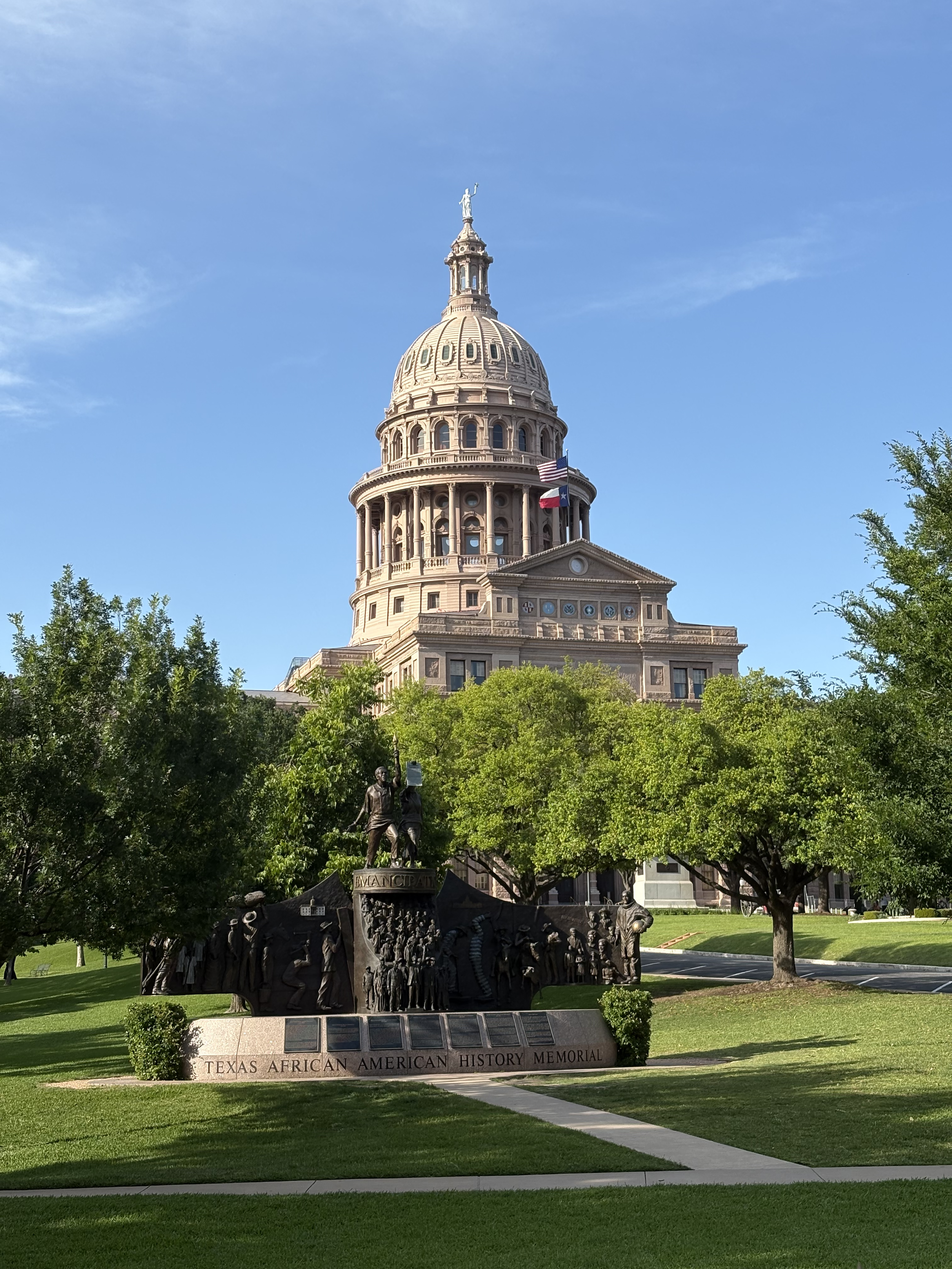 The Texas State Capitol building, seen through green trees with a bright blue sky in the background. The dome is topped with a statue and an American flag and Texas flag are hanging from the building.