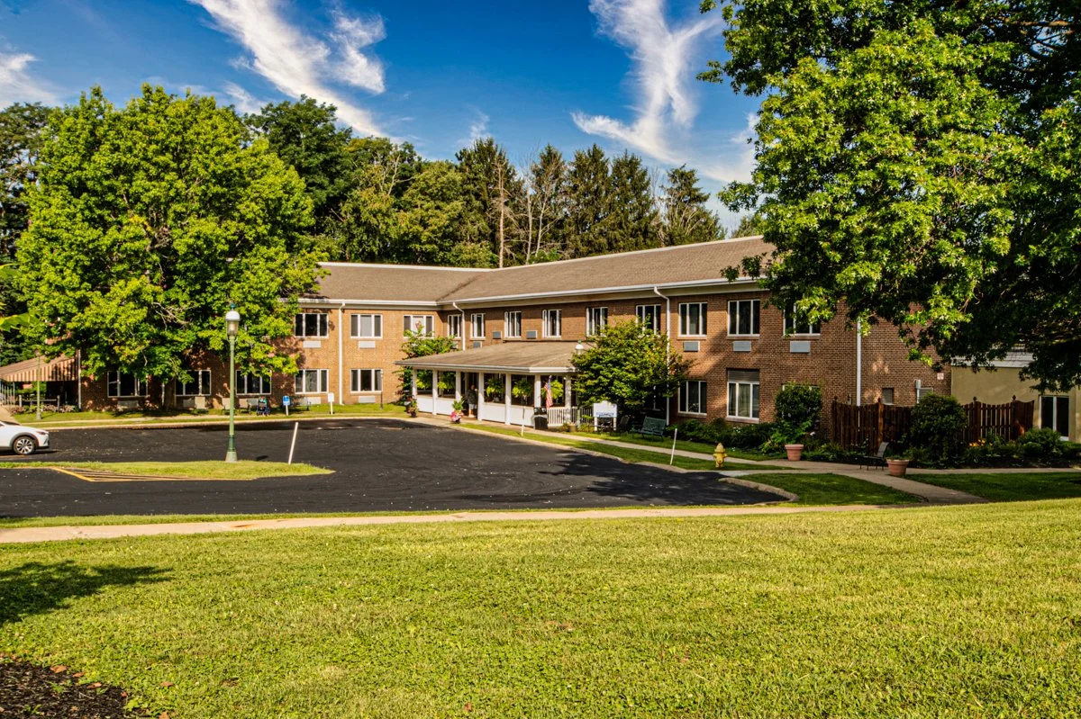 A multi-story brick apartment building with a porch, surrounded by green trees and a well-maintained lawn, under a partly cloudy sky.