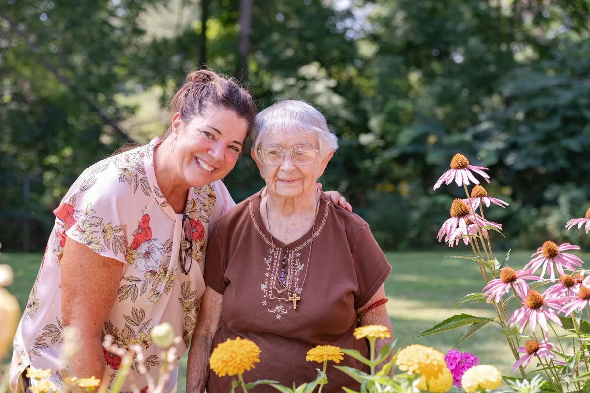 A woman and an elderly woman smiling in a garden with flowers and trees in the background.