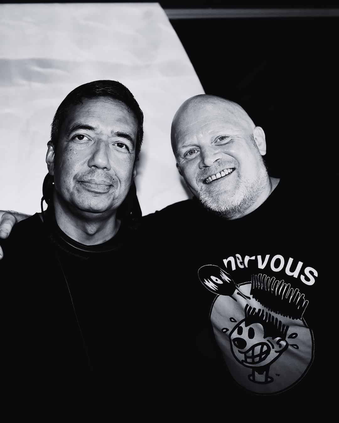 Two men taking a selfie together, one with dark hair and the other with a bald head and beard, in black and white. The man with the beard is wearing a T-shirt with the word 'nervous' and a cartoon character on it.