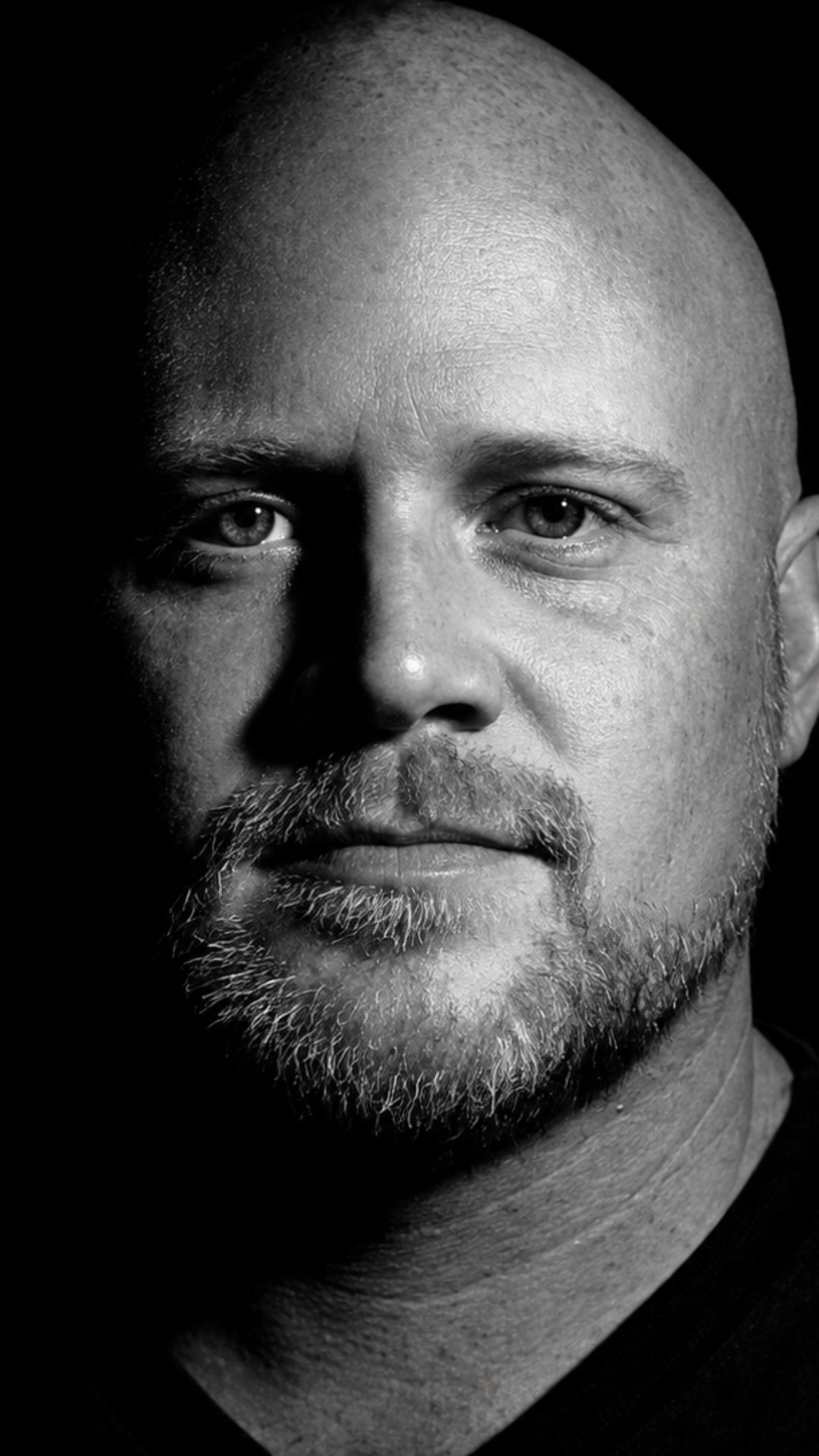 Close-up black and white portrait of Merah with a bald head, light beard, and piercing eyes, looking directly at the camera.