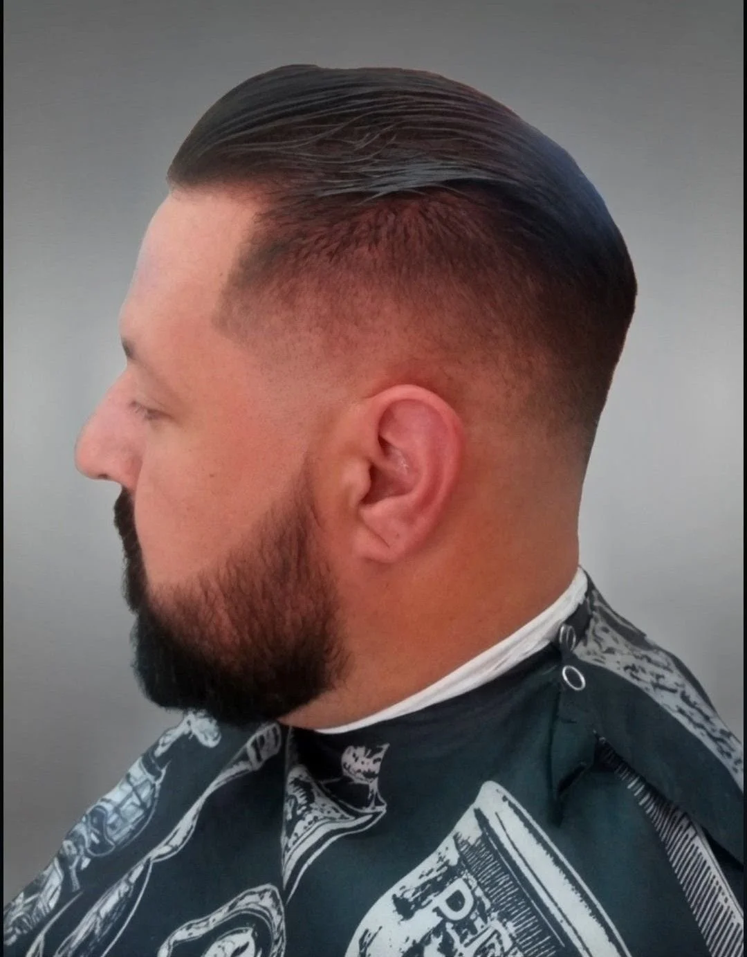 Side profile of a man with a fresh fade haircut, neatly combed dark hair, full beard, and wearing a barber cape with graffiti-like designs.