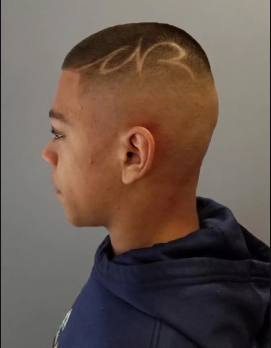 Side profile of a young man with a fresh haircut featuring a design shaved into the side, wearing a dark hoodie.