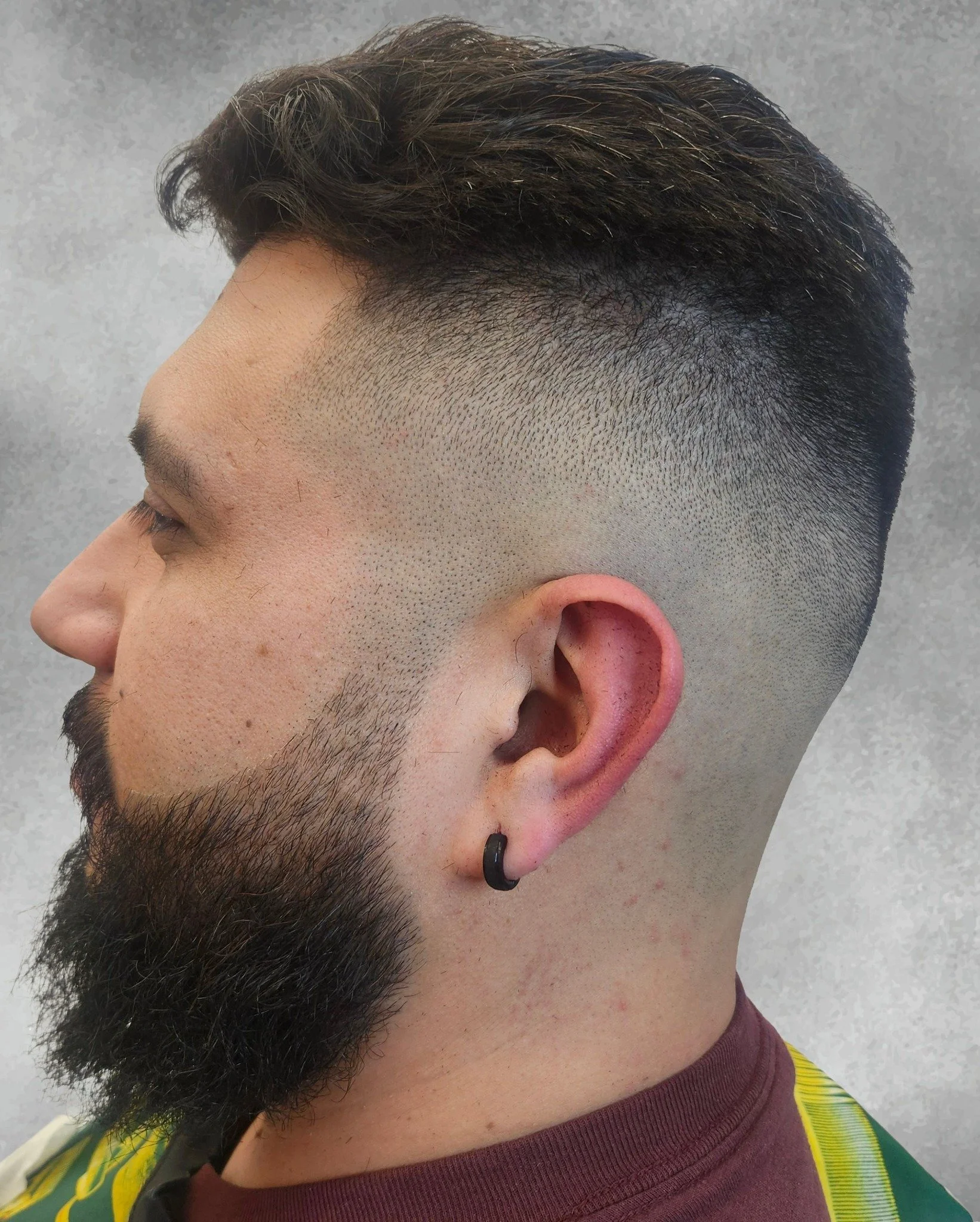 Side profile of a man with dark hair done in an undercut hairstyle, beard, and mustache, wearing a black earring and colorful clothing.