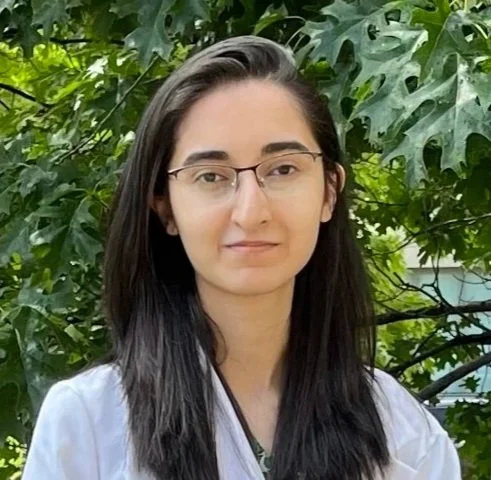 A young woman with long dark hair, glasses, and light skin stands outdoors in front of green leafy trees, wearing a white top.