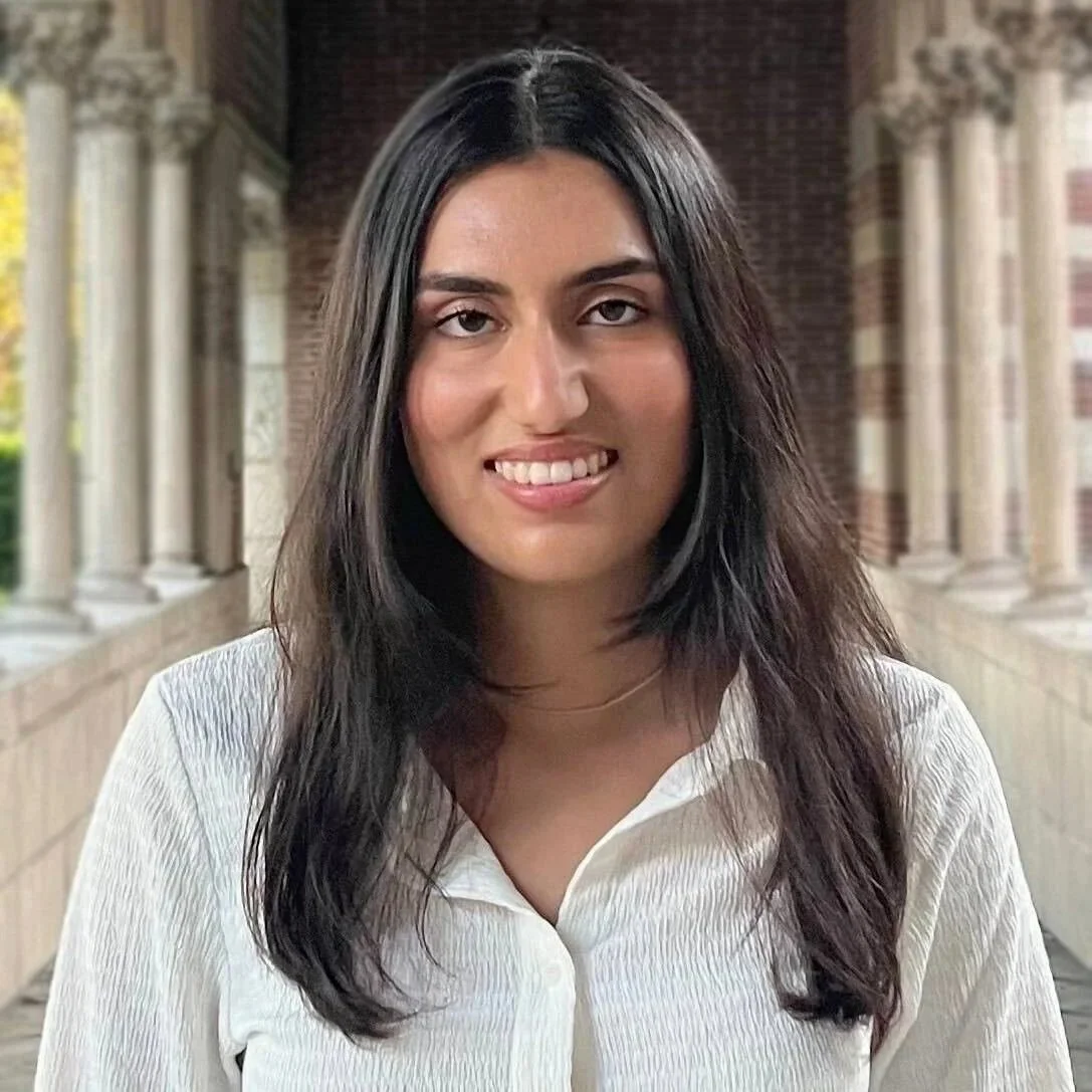 A woman with shoulder-length dark hair, wearing a white top, smiling and sticking out her tongue slightly, standing outdoors with columns and a brick wall in the background.