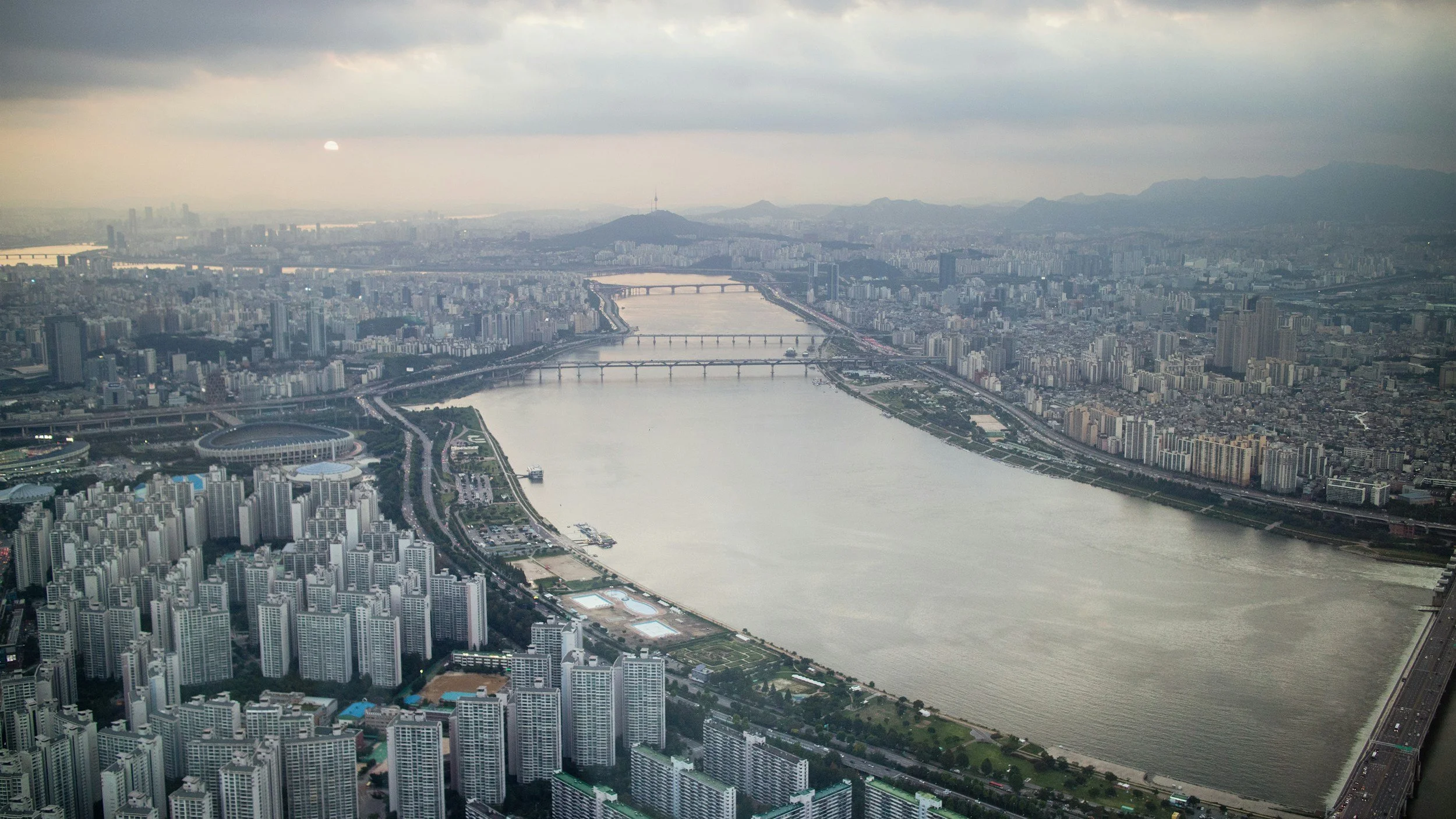 Aerial view of a large city with numerous high-rise buildings, a river with multiple bridges, and mountains in the background during cloudy weather.