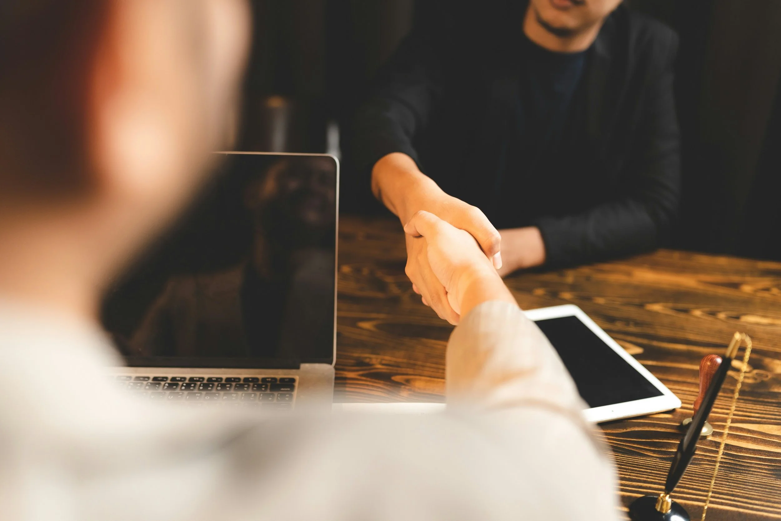 Two people shaking hands on a wooden table with laptops and tablets around them, indicating a business meeting or agreement.