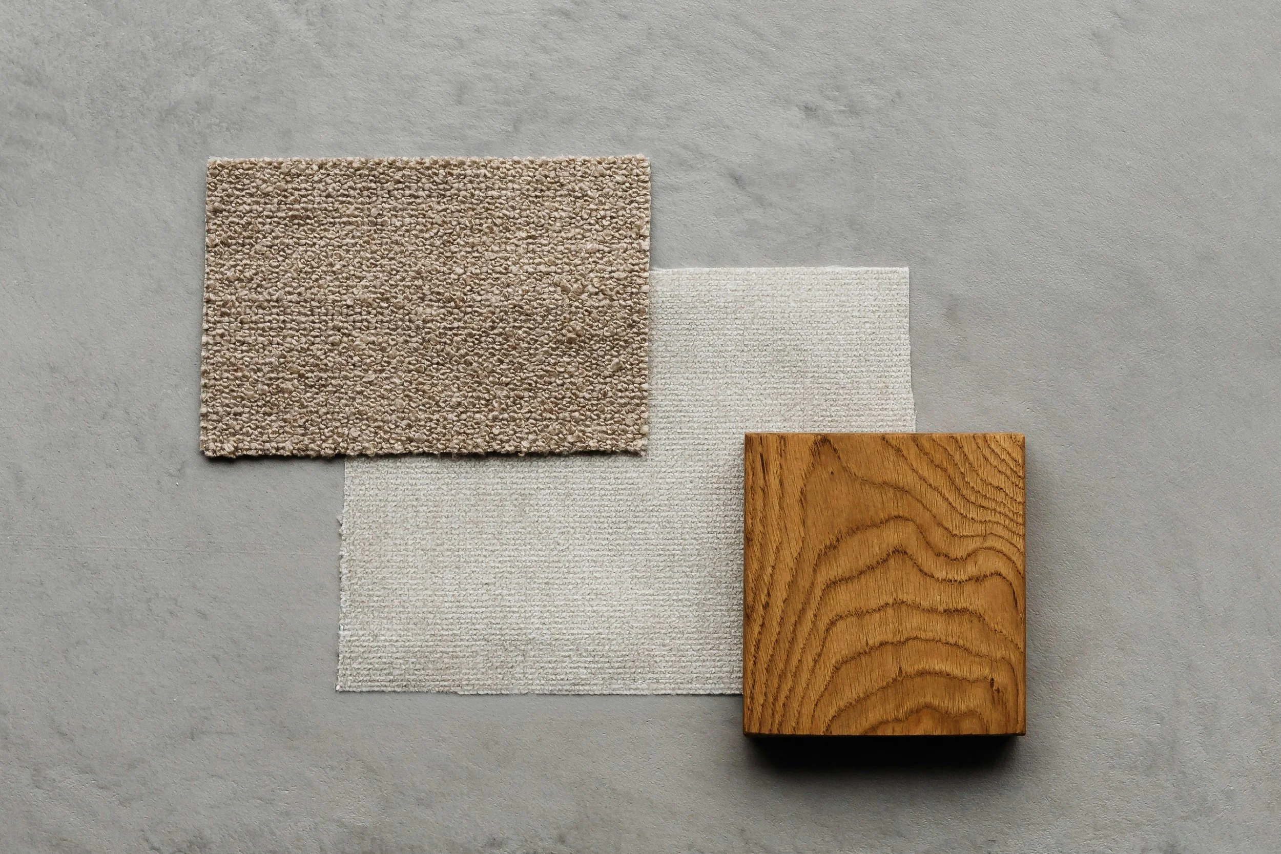 An arrangement of four different material samples on a gray background, including a beige carpet swatch, a light-colored woven fabric, a piece of wooden panel, and a smooth gray surface.