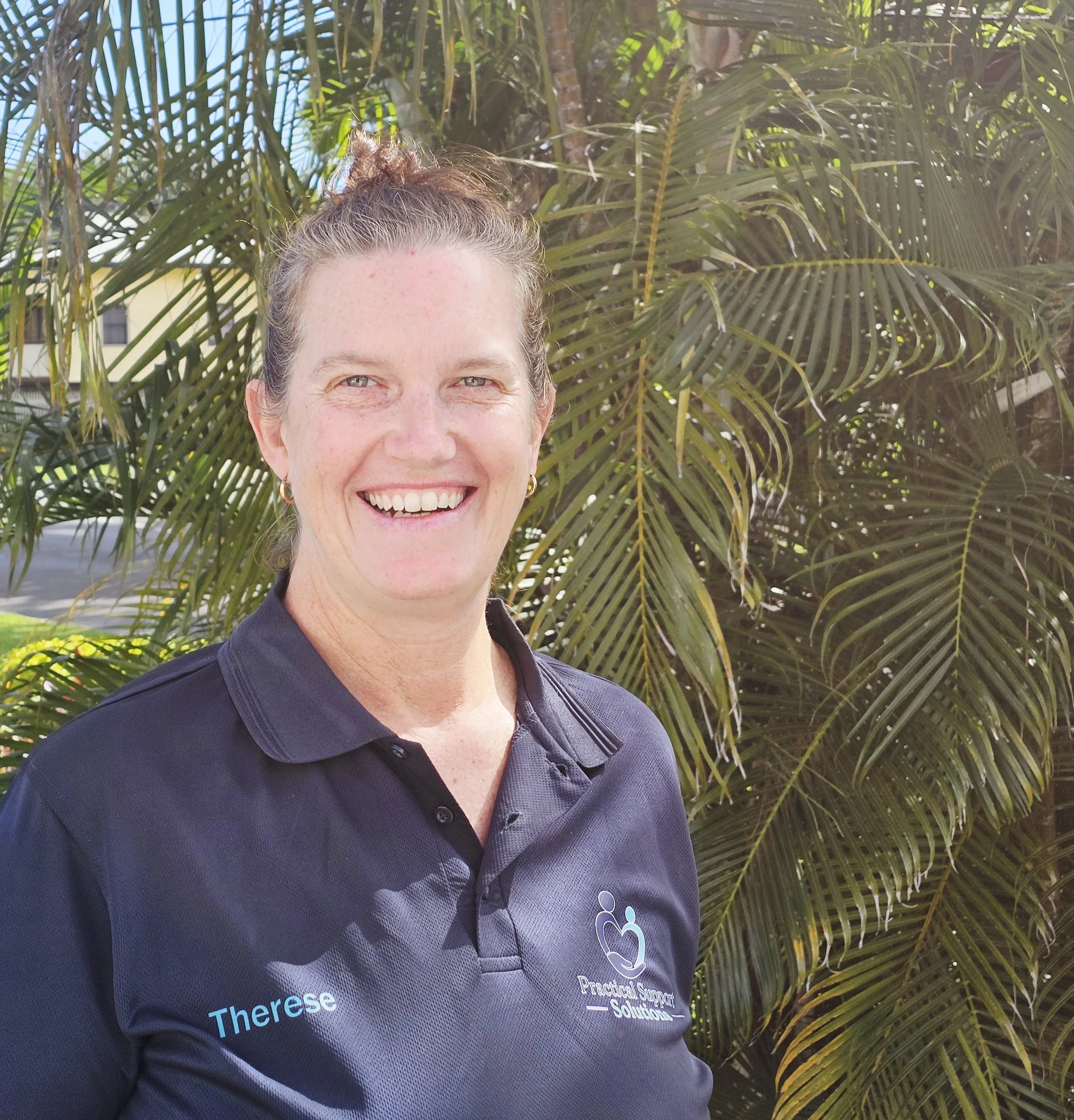A woman smiling outdoors in front of palm trees, wearing a black polo shirt with Nike logo and text on it.