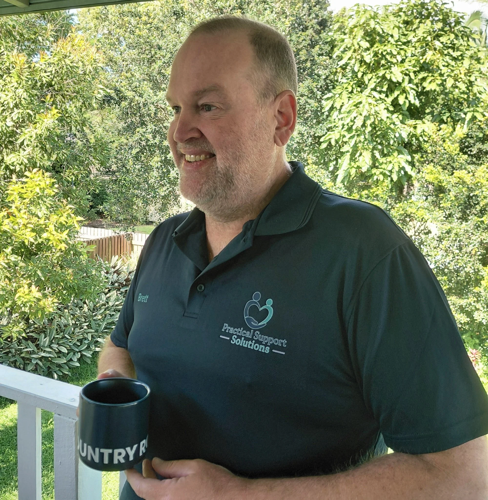 A man holding a black mug with the words 'UNTRY R' and a partial word visible, standing outdoors in front of lush green trees and bushes, smiling and wearing a black shirt with a logo and text on it.