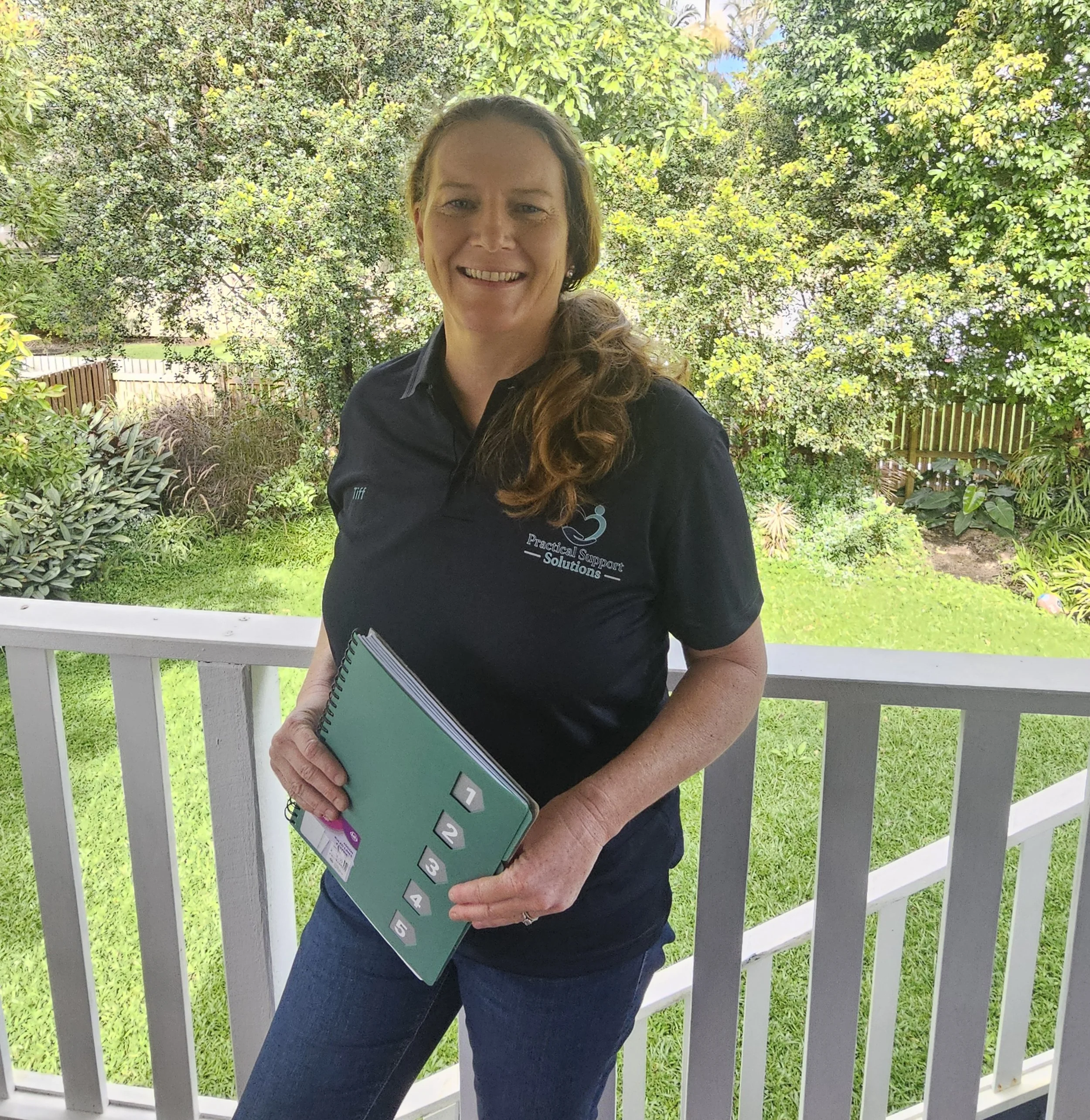 A woman standing outdoors on a porch holding a blueprint, smiling at the camera, surrounded by green trees and plants, wearing a navy shirt with logo and jeans.