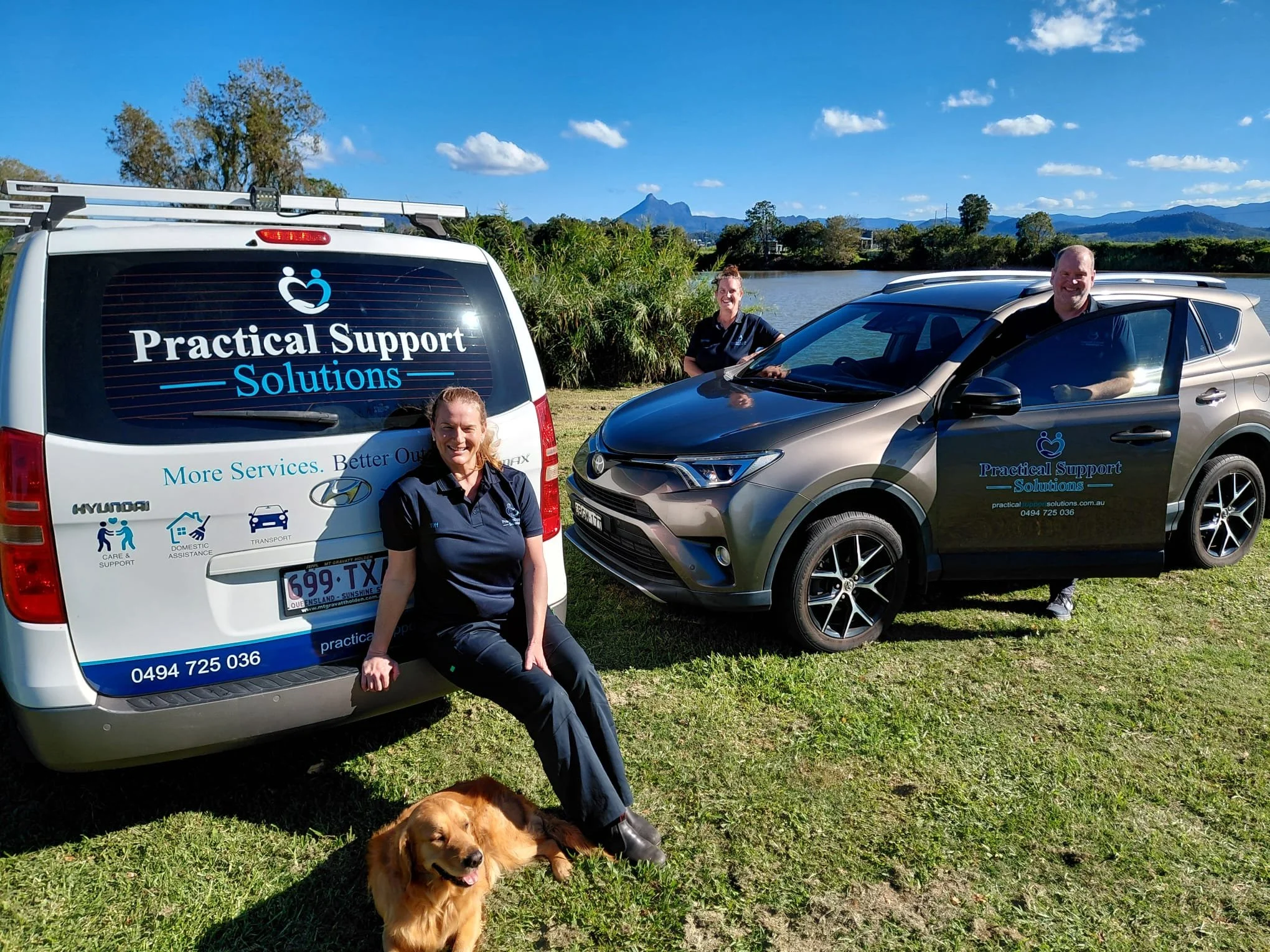 A group of three smiling people and a dog with a landscape of mountains, trees, and a river in the background. They are outdoors next to vehicles with company branding for Practical Support Solutions, and the sky is clear with a few clouds.